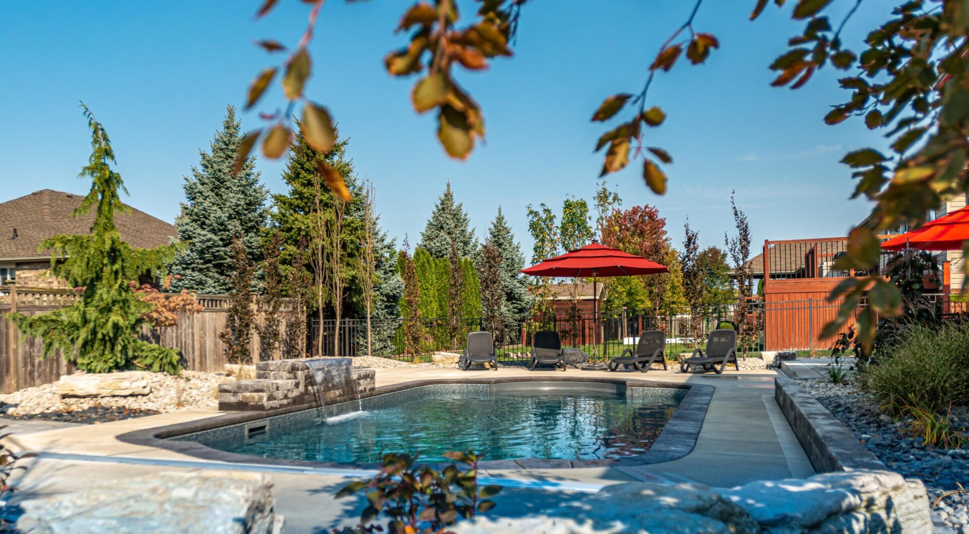 A serene backyard with an inviting swimming pool, surrounded by loungers and red umbrellas, set against a backdrop of trees and a clear blue sky.