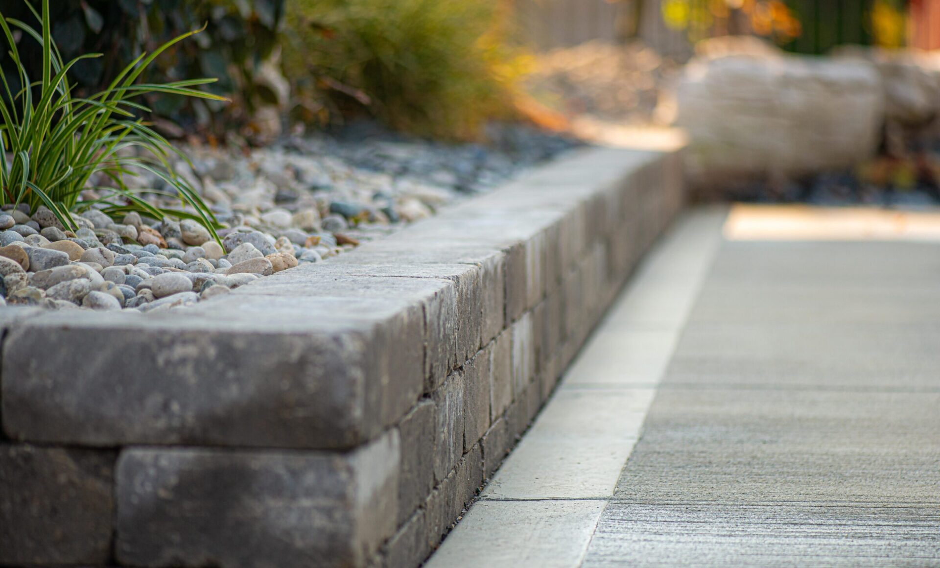 A neatly composed image of a landscaped garden edge, featuring a low brick retaining wall, a gravel bed, some green plants, and a concrete pathway.