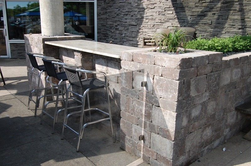Outdoor seating area with stone bar counter, four metal bar stools, and plants, next to a structured stone pillar with a clear sky in the background.