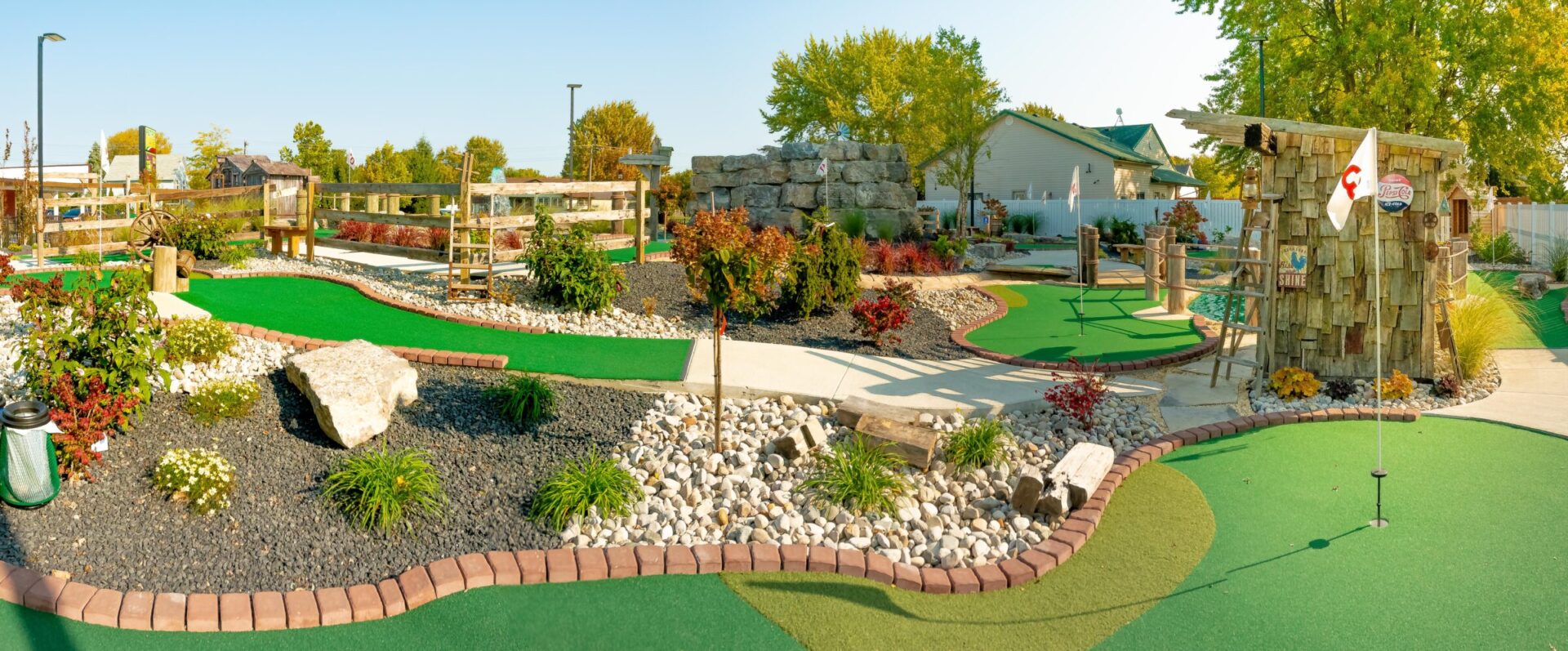 This panoramic image shows a vibrant outdoor miniature golf course with lush landscaping, various obstacles, rocks, and a rustic-themed watermill hut.