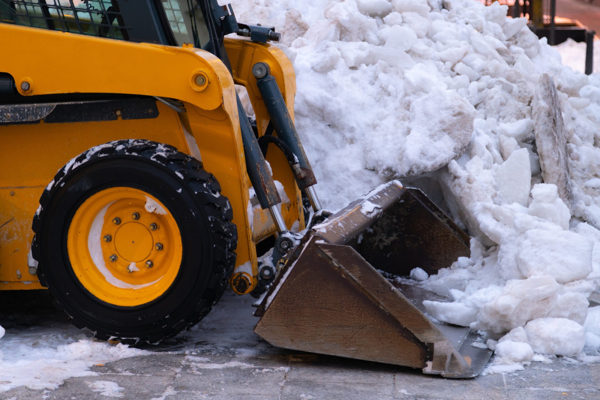A yellow skid steer loader with a bucket attachment is clearing a large pile of dirty snow from a paved surface in a cold environment.