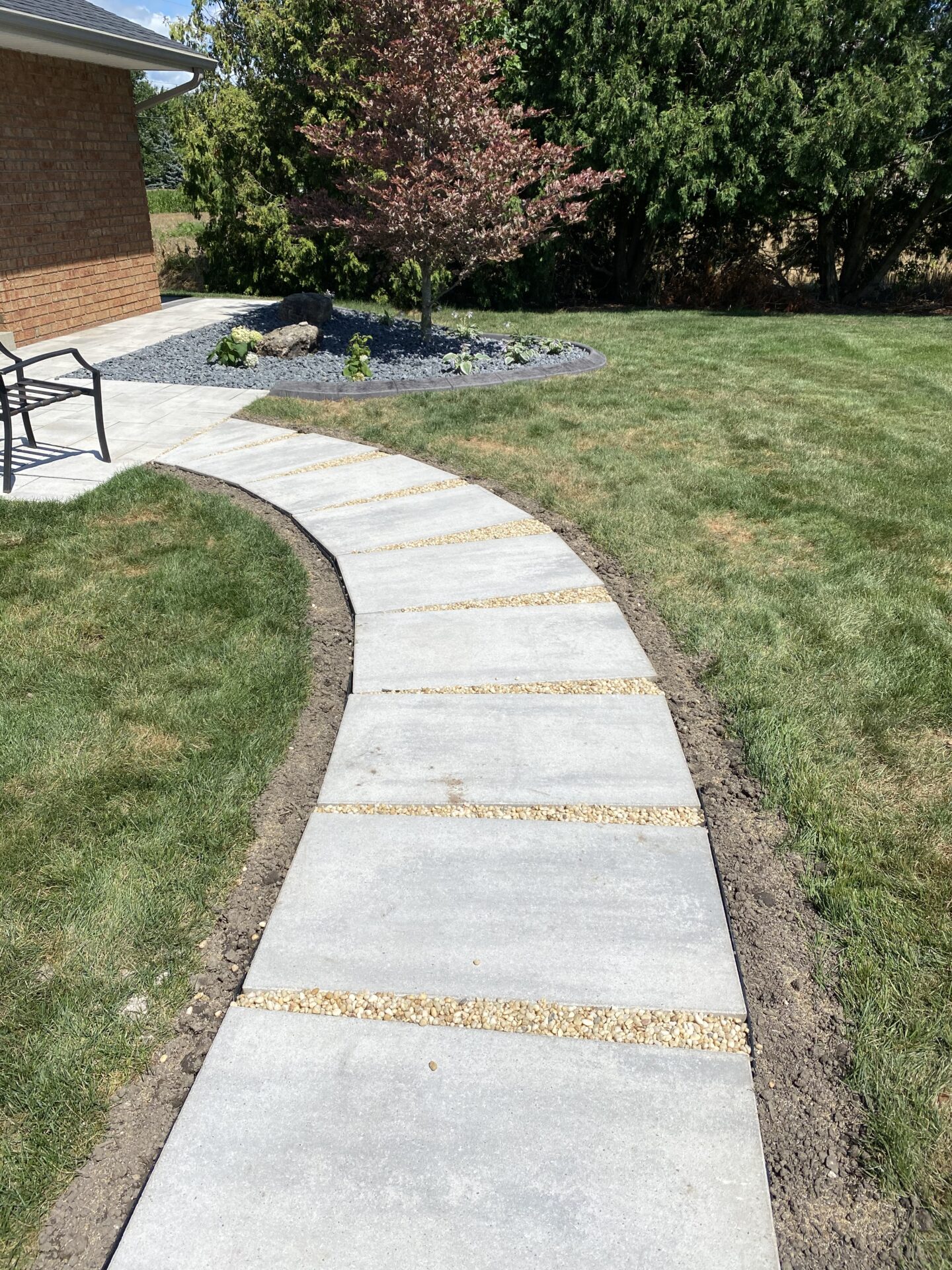 This is a well-manicured garden pathway with rectangular stepping stones and gravel accents, leading towards a patio area with a seating arrangement.