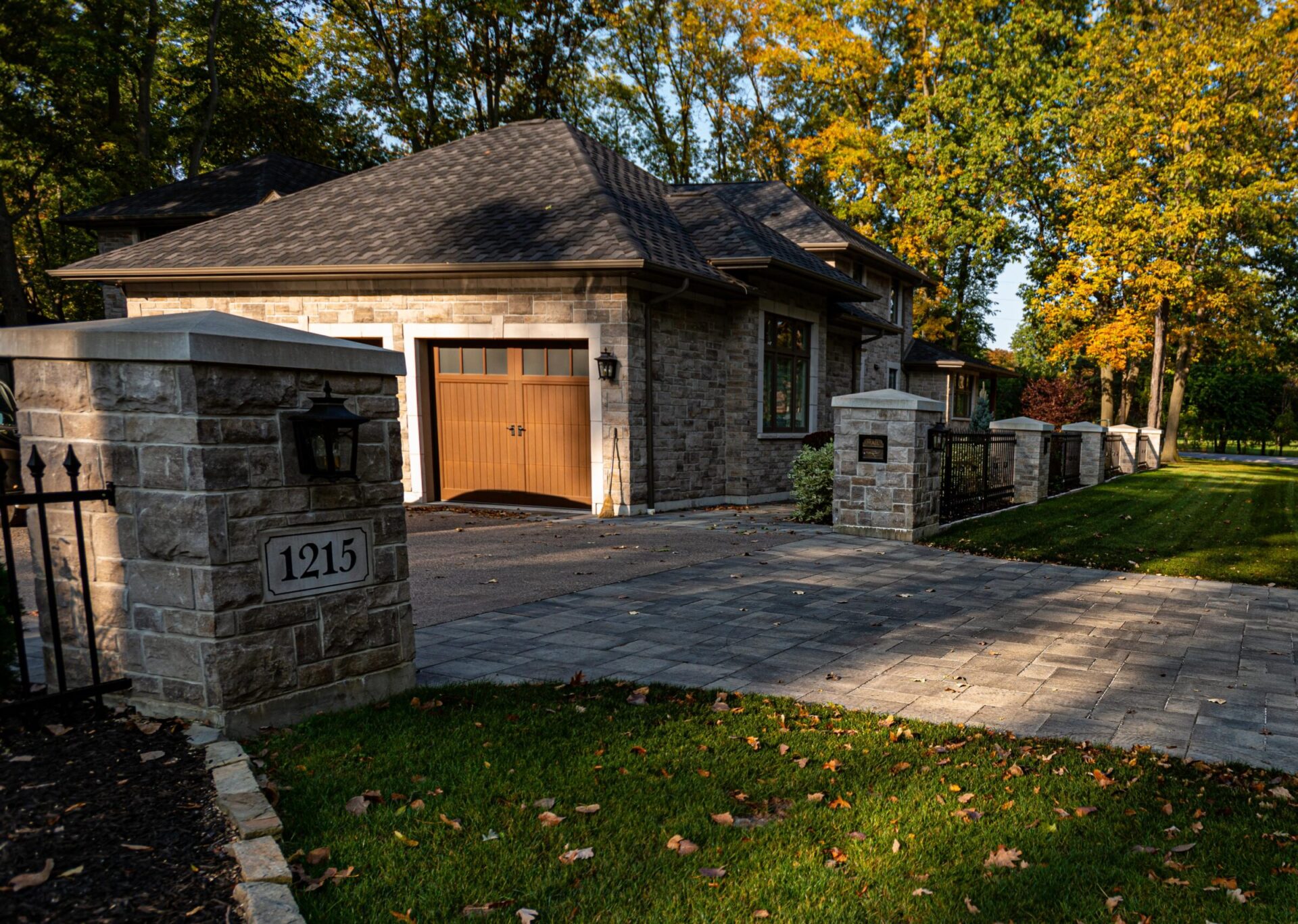 A stone house with a wooden garage door surrounded by a stone fence, lantern, and a well-manicured lawn under autumn trees with golden leaves.