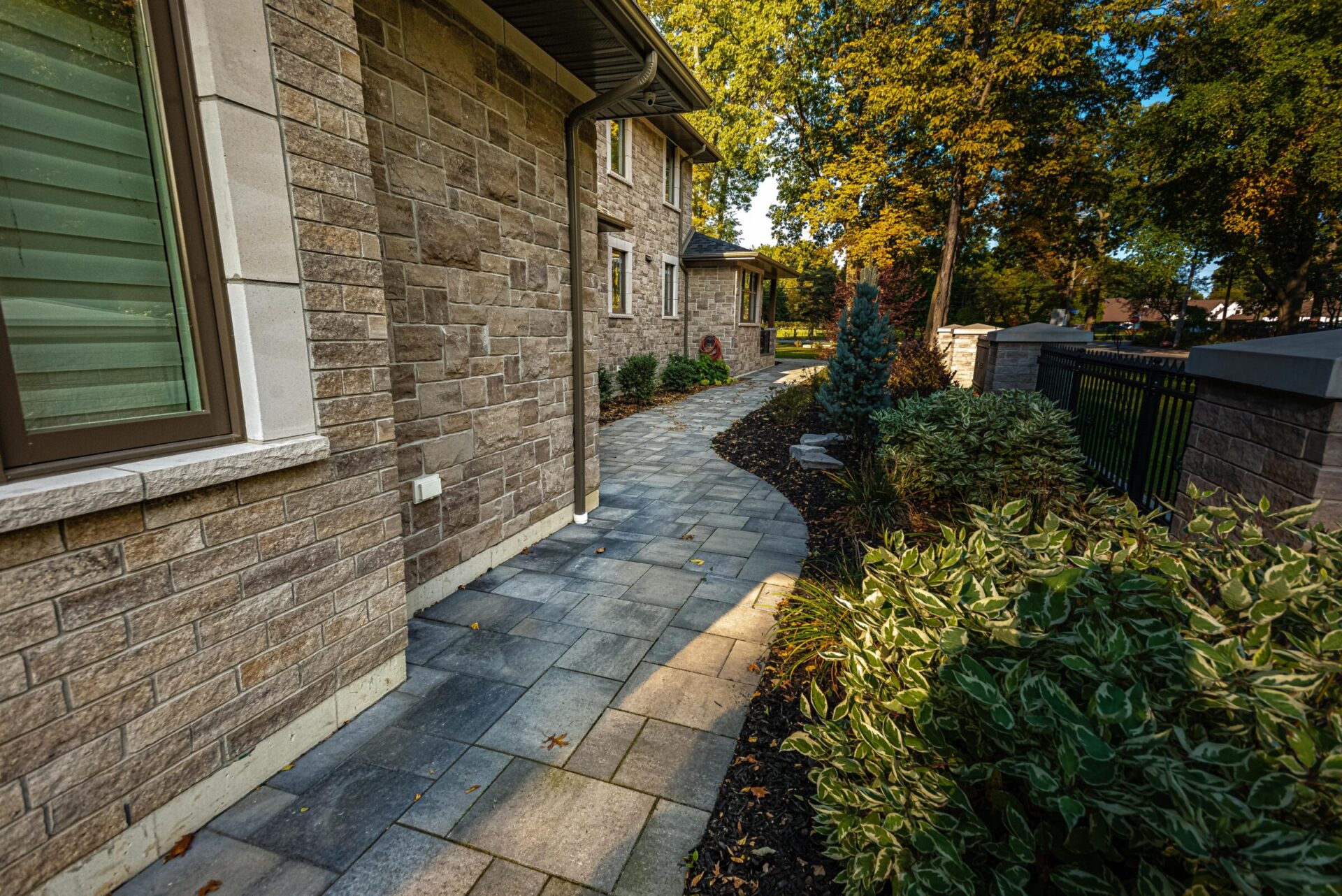 A curved stone walkway leads to a two-story residential house with brick exterior, flanked by lush green shrubbery and mature trees under clear skies.