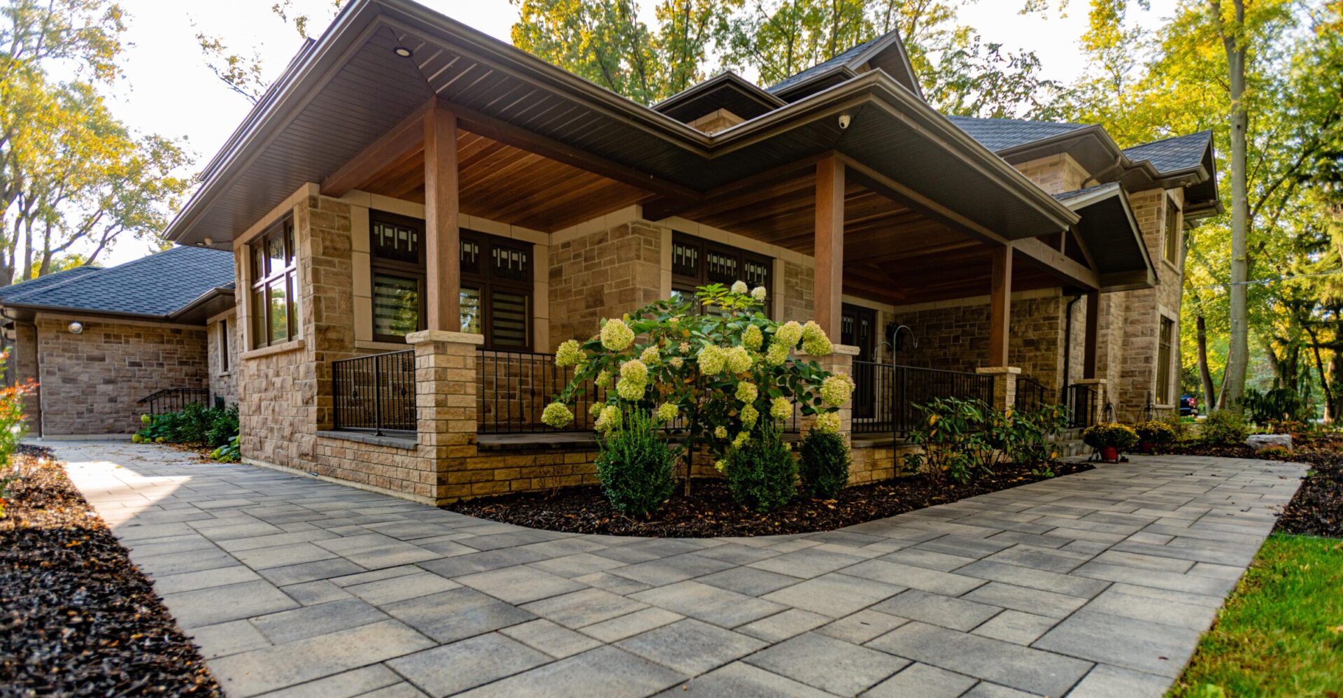 This is an image of a modern house with a large porch, stone pillars, landscaped garden, hydrangea bushes, and a paved walkway, surrounded by trees.