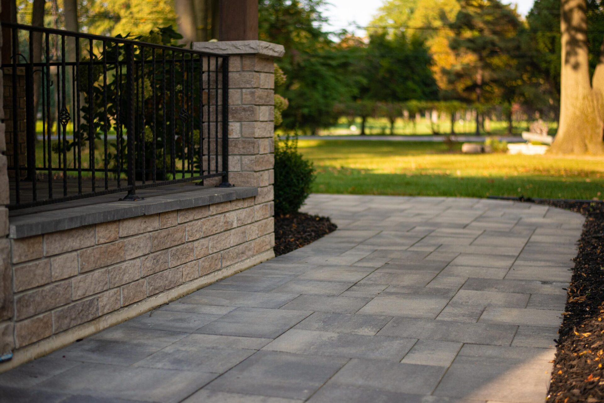 This image shows a paved pathway leading to an iron fence and brick column, with a garden and trees in the soft-focused background at dusk.