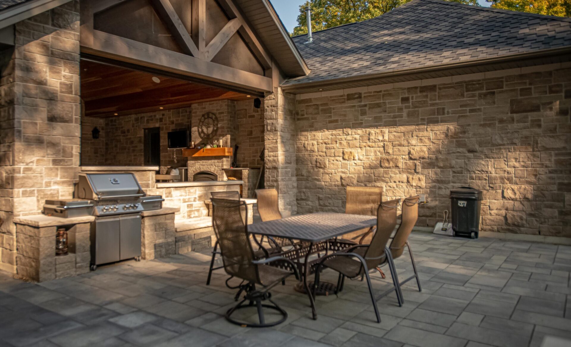 An outdoor patio with stone walls features a dining set, barbecue grill, under a pergola with a wooden ceiling on a sunny day.