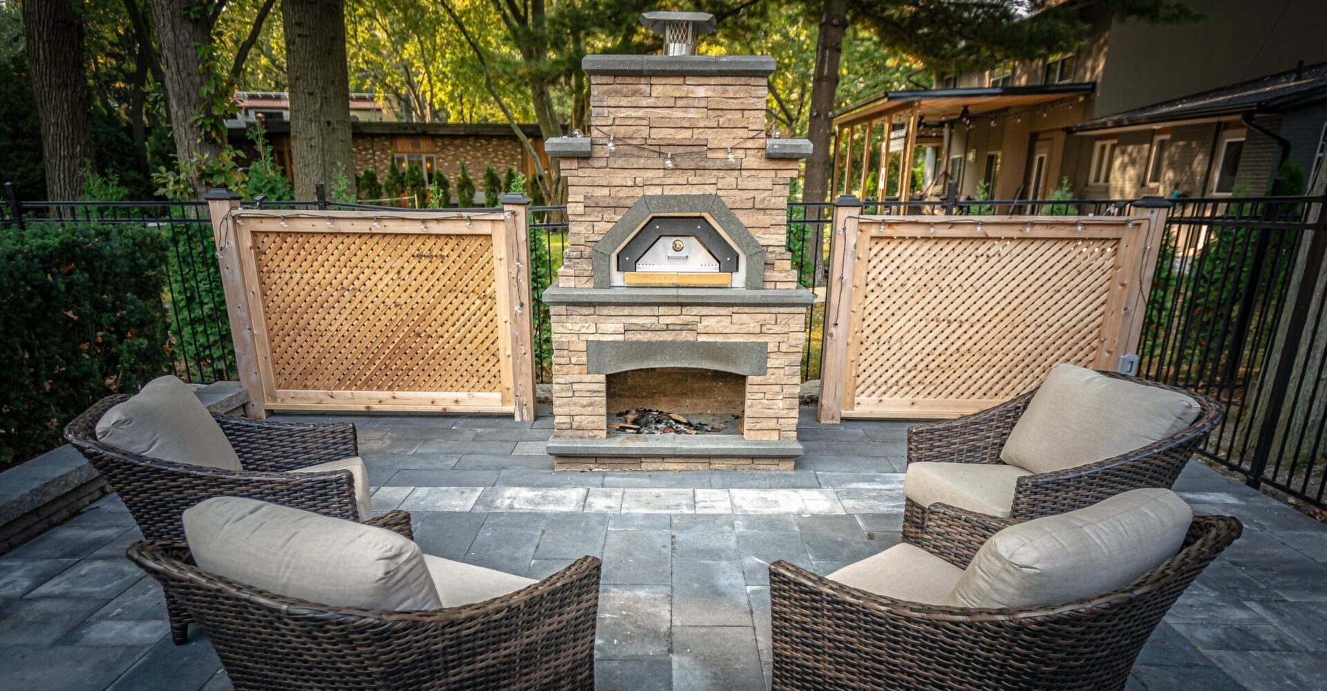 An outdoor patio features a stone pizza oven, wicker chairs with cushions, a wooden lattice privacy screen, and a patterned stone floor surrounded by trees.