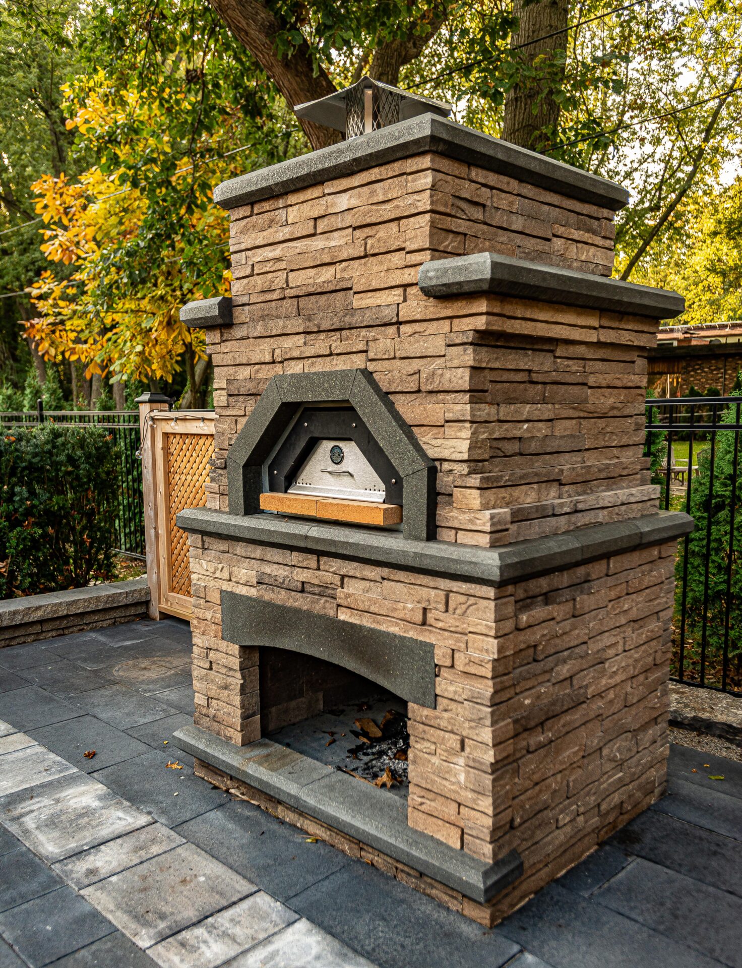An outdoor stone pizza oven with a chimney stands on a patterned pavement, surrounded by a wooden fence and trees with autumn leaves.