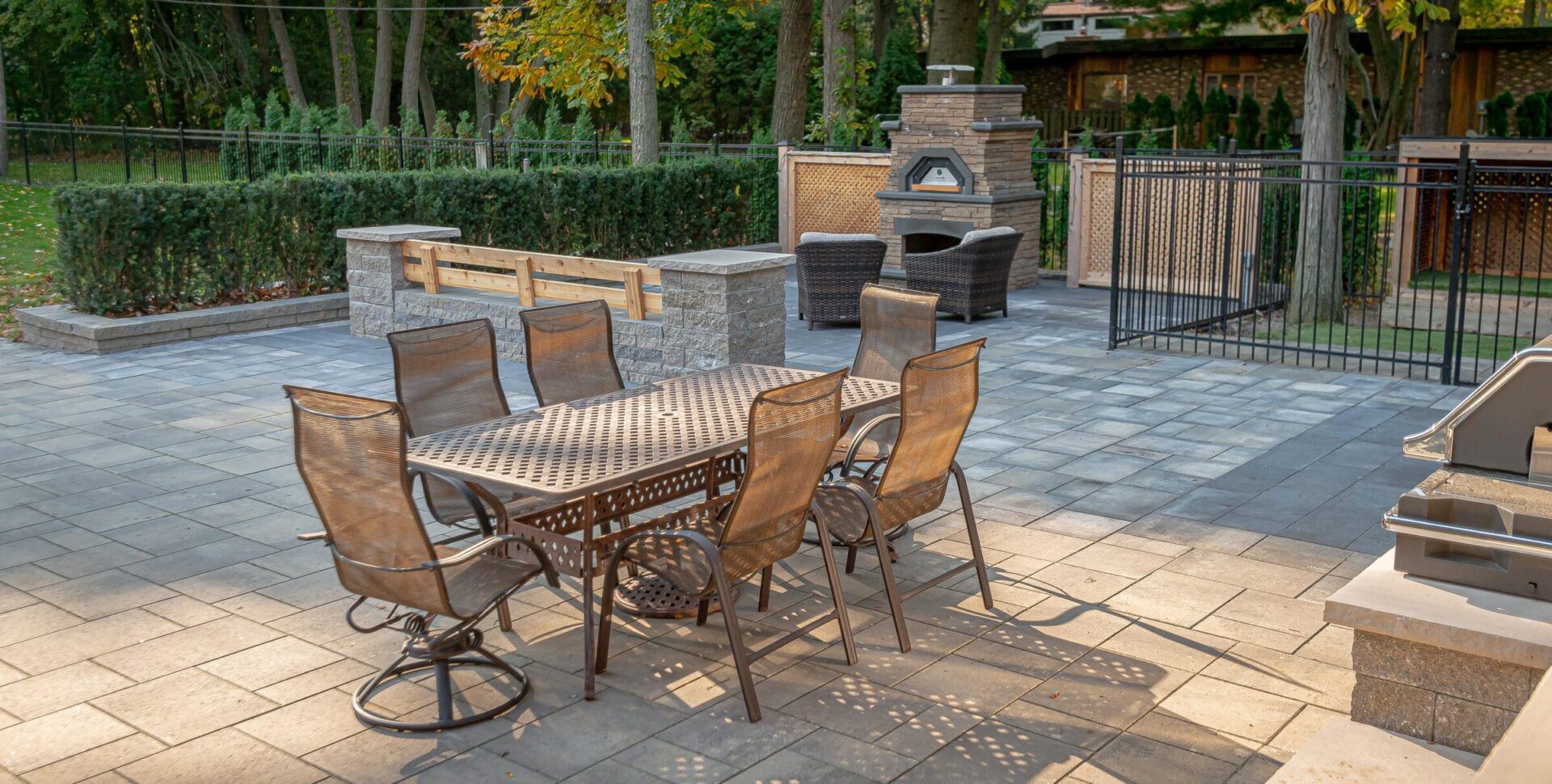 An outdoor patio with a dining table, chairs, stone benches, a pizza oven, and a grill set in a landscaped yard enclosed by a metal fence.