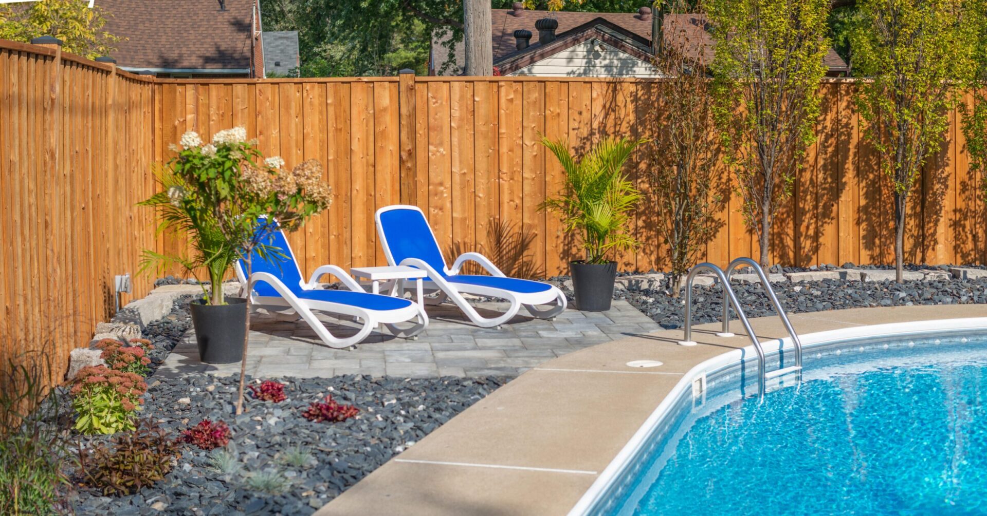 A backyard with a swimming pool, blue lounge chairs, landscaped with rocks and plants, surrounded by a tall wooden fence, under a clear sky.