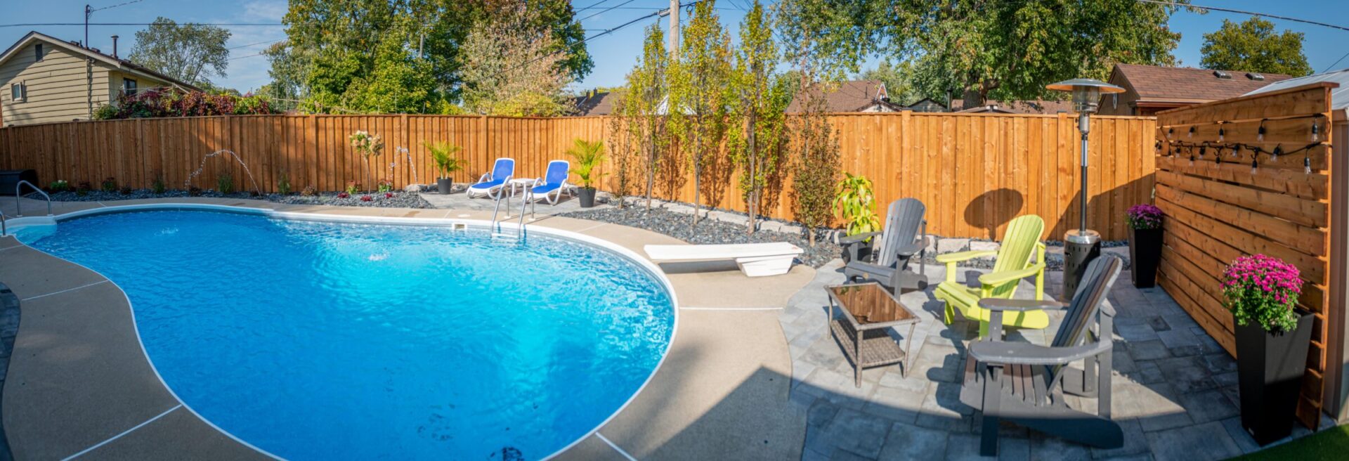 A panoramic view of a backyard with a curvy swimming pool, sun loungers, patio furniture, plants, a wooden fence, and string lights.