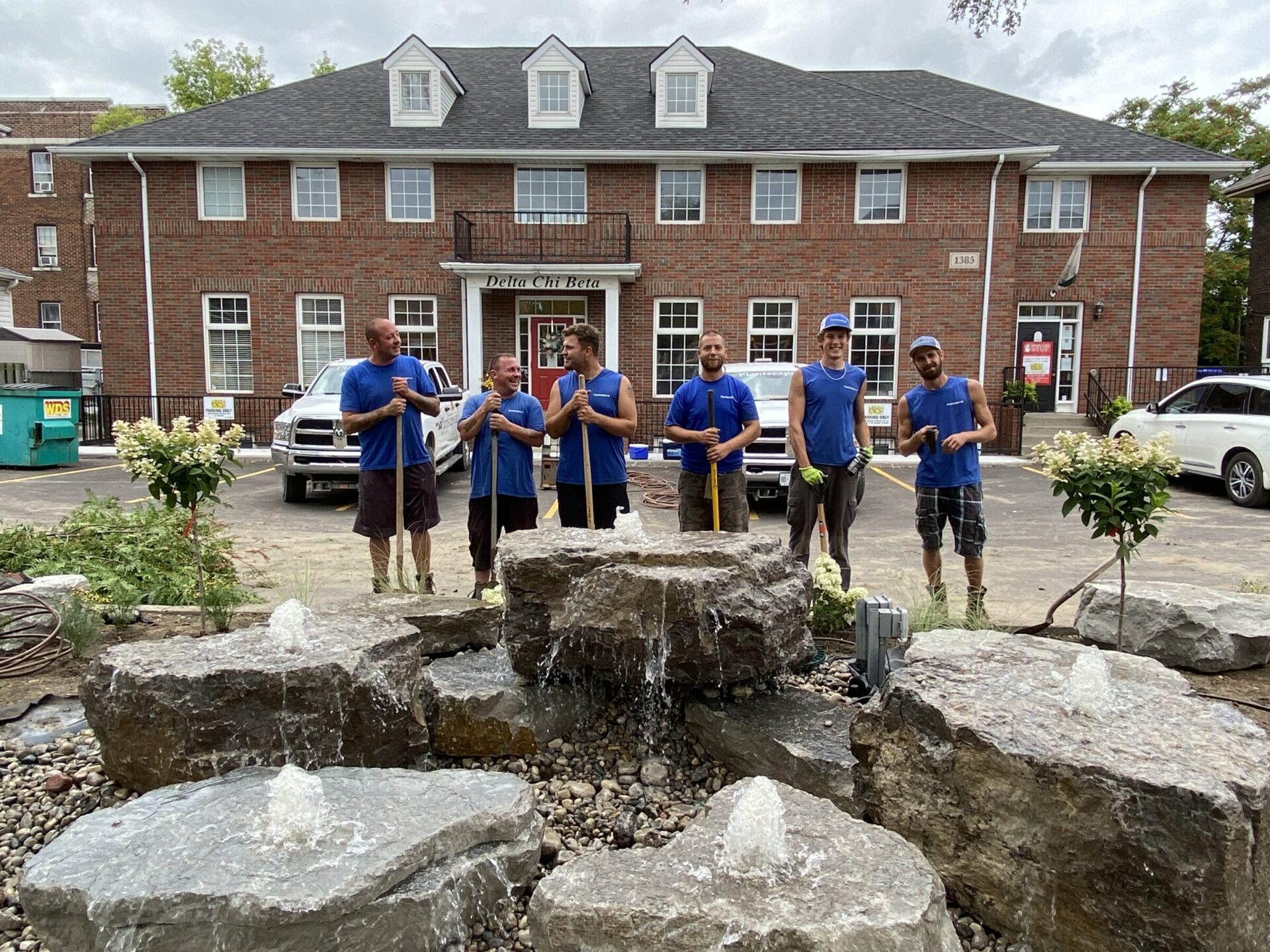 Five people stand near a water feature composed of rocks and cascading water. They wear matching blue shirts, potentially indicating a team or work crew.