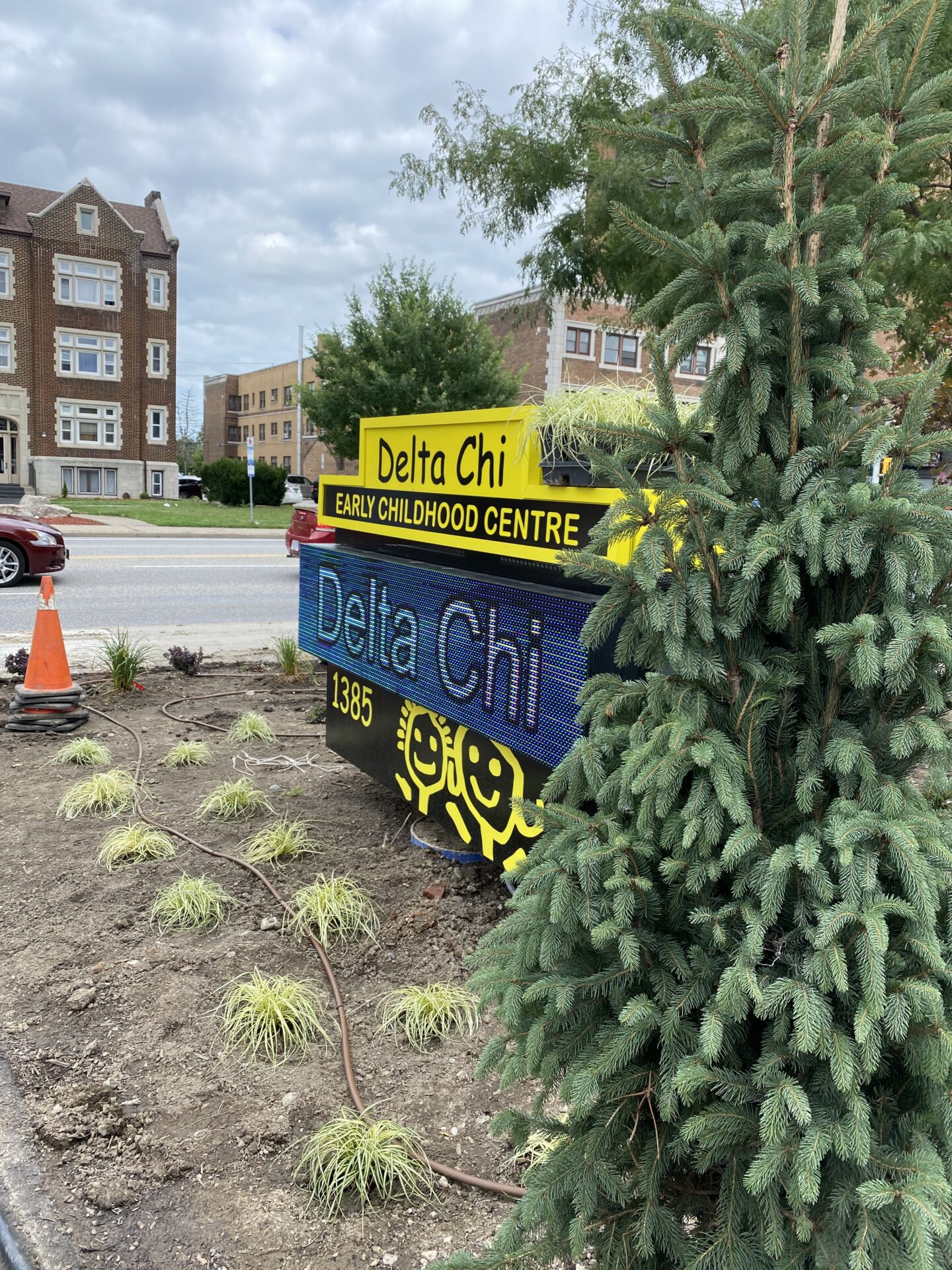 A digital sign for Delta Chi Early Childhood Centre is displayed beside a tree with landscaping. Tall buildings and a car are in the background.
