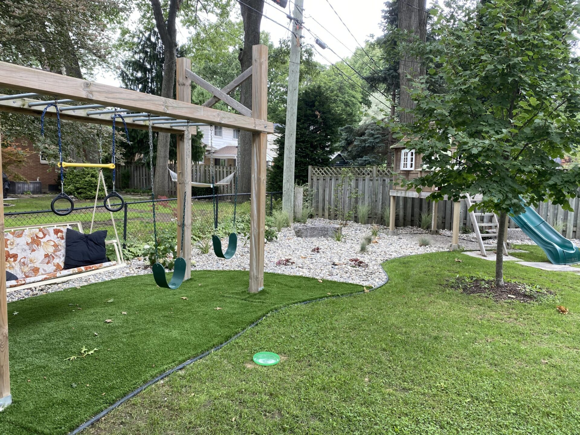 A backyard with artificial turf, a wooden swing set, a slide, trees, rocks, a bench with cushions, and a small, decorative fence.