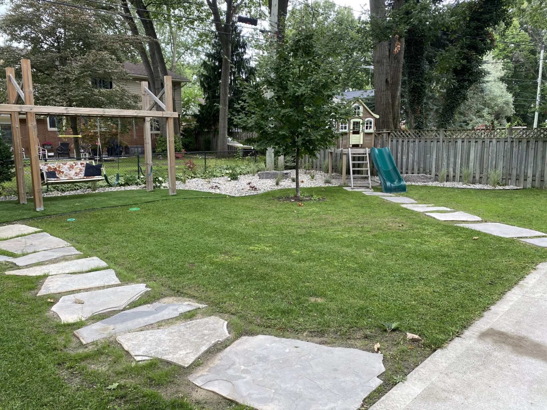 A backyard with a green lawn featuring a swing set, a playhouse on stilts with a slide, stepping stones, trees, and a wooden fence.