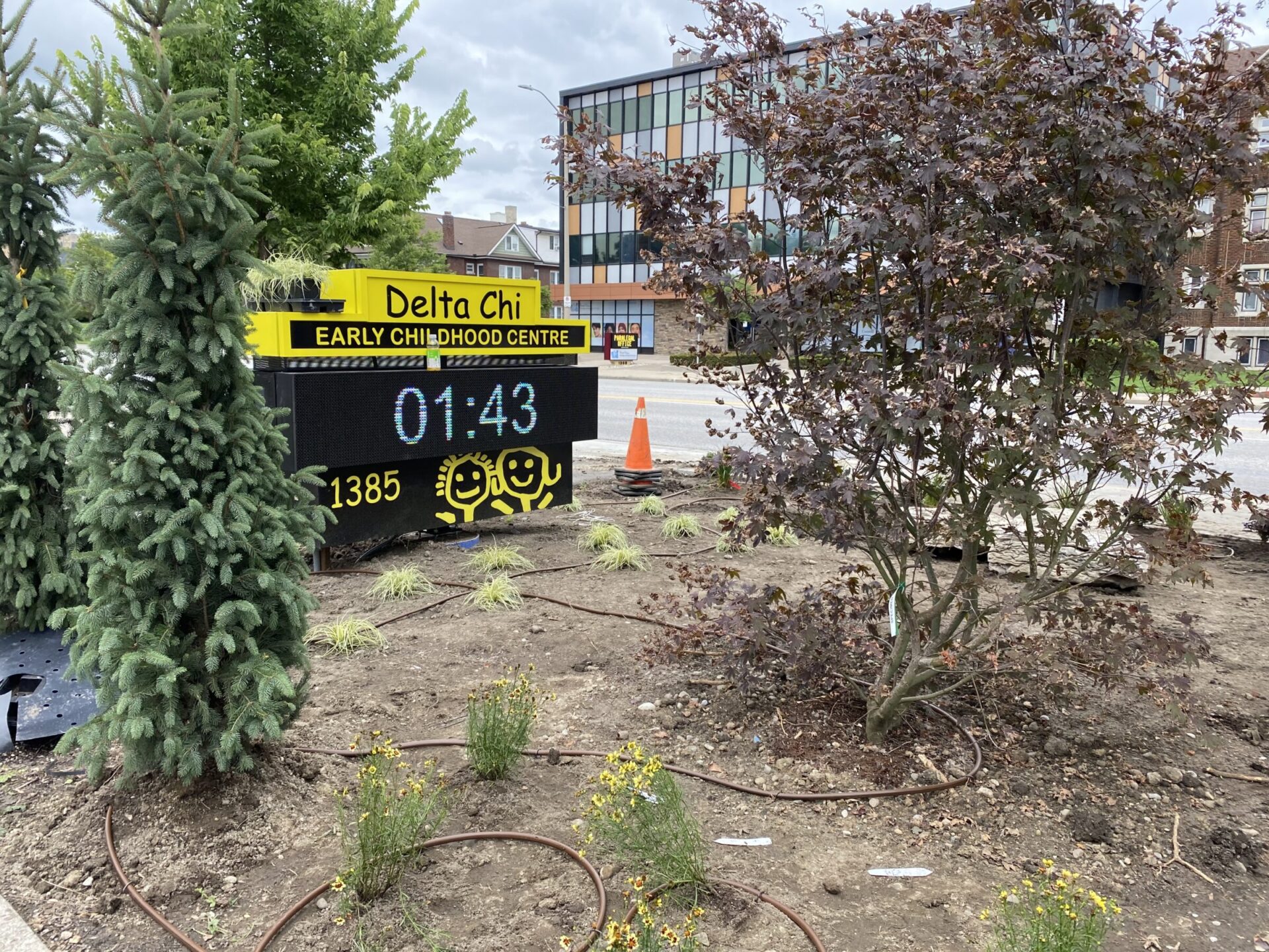 The image shows a landscaped area with a digital sign for Delta Chi Early Childhood Centre, displaying the time 01:43 with happy face emojis and vegetation around.