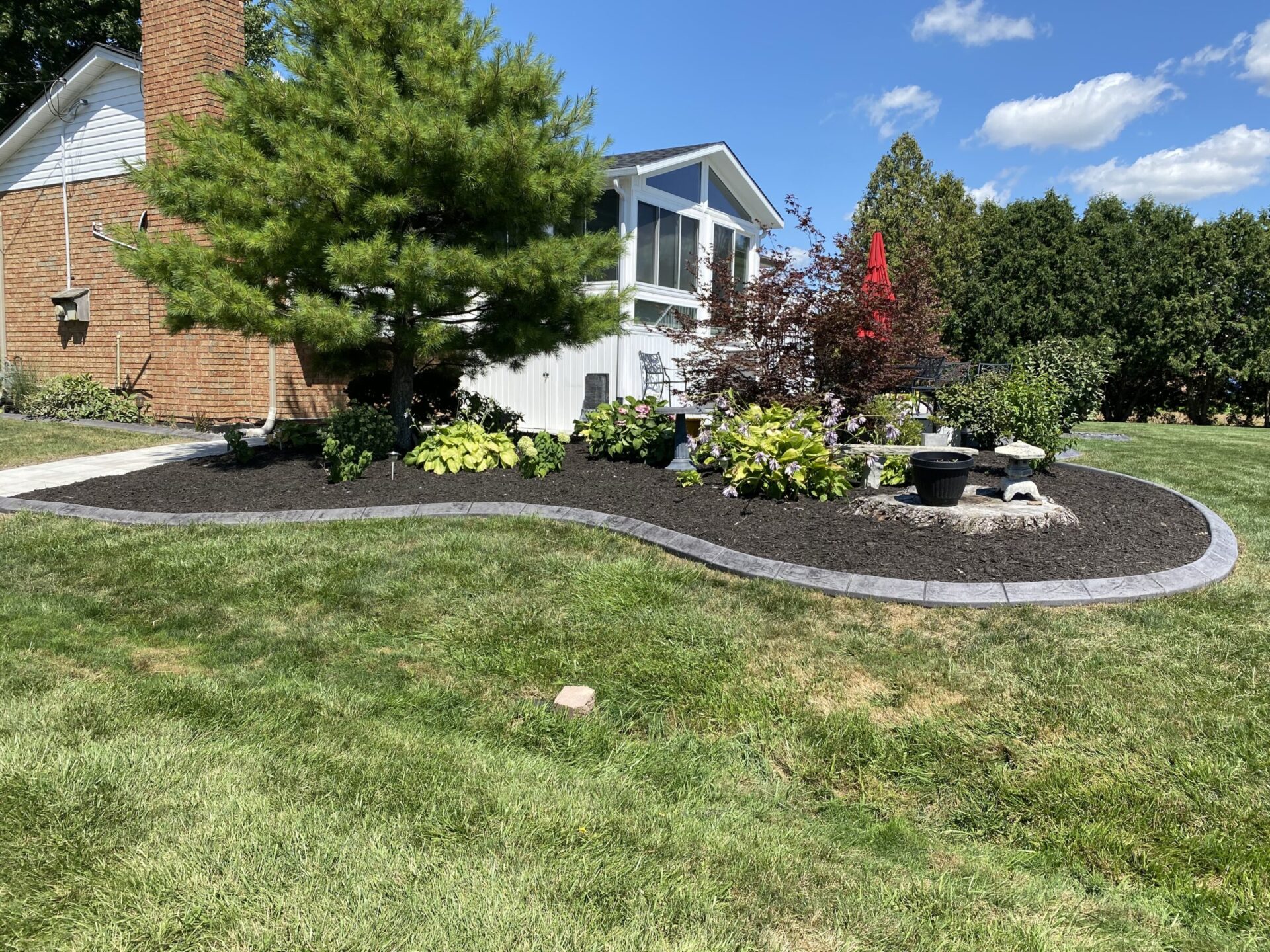 A neatly manicured garden with a curved mulch bed, assorted plants, a tree, and a red wind spinner in front of a home.