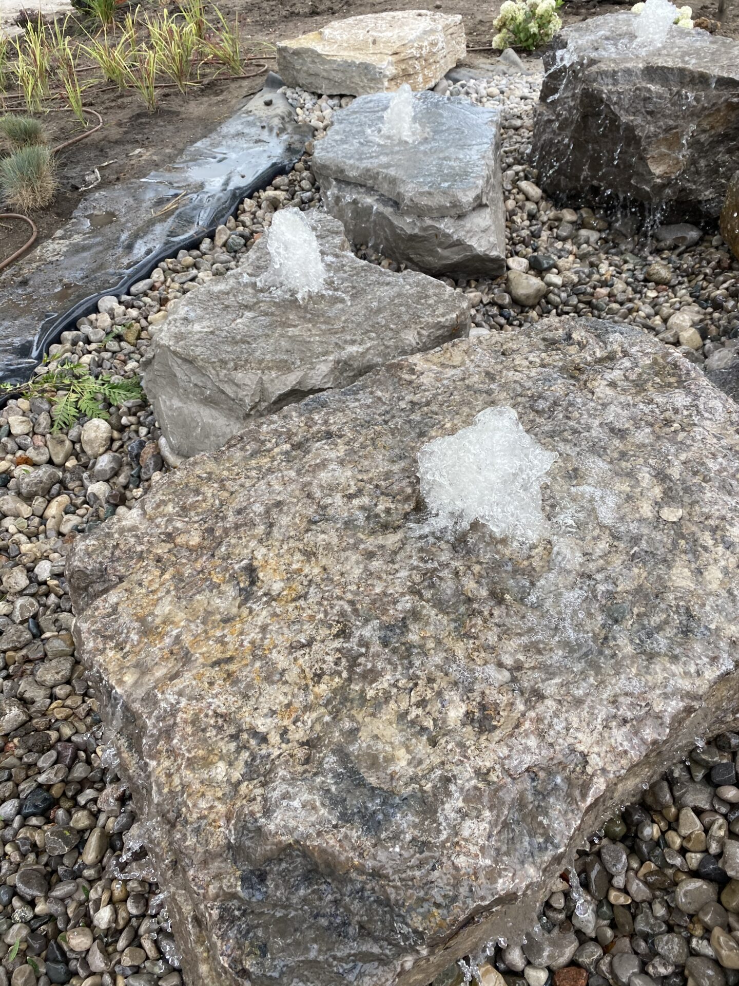 This image features a rocky landscape with water splashing on large stones, surrounded by smaller pebbles and vegetation, possibly part of a garden feature.