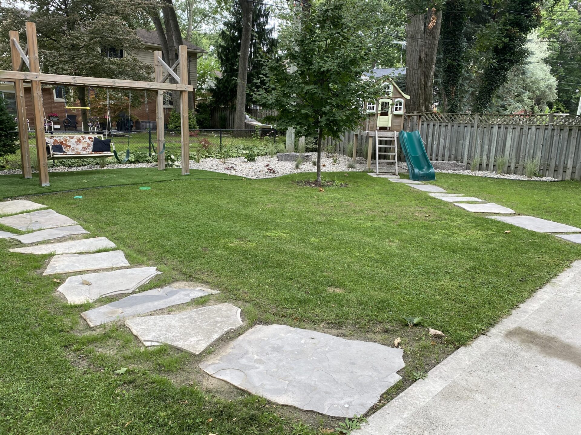 A residential backyard with stone pathway, green grass, a swing set, playhouse, and slide, surrounded by trees and a wooden fence.