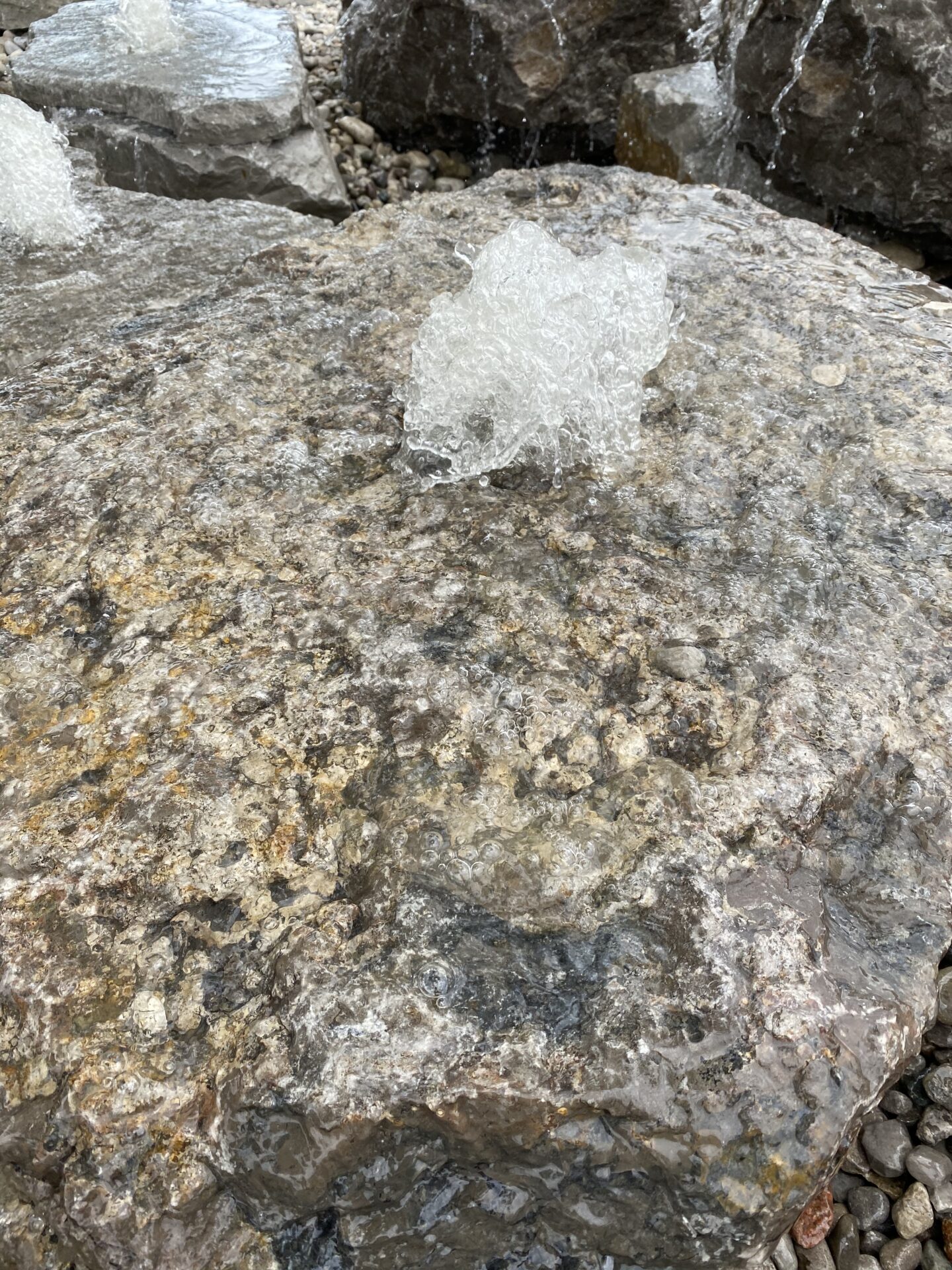 The image shows water bubbling over a large rock, possibly in a stream or fountain, with additional rocks and flowing water visible in the background.