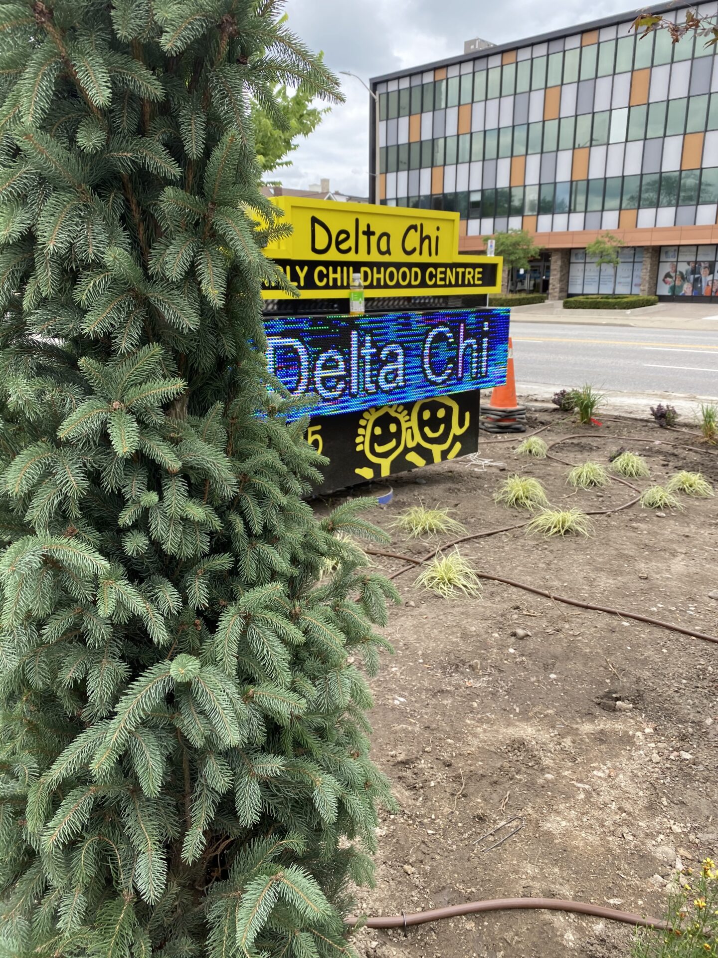 This image shows a sign for Delta Chi Early Childhood Centre, with LED display and smiley faces, in front of a building beside a conifer tree.