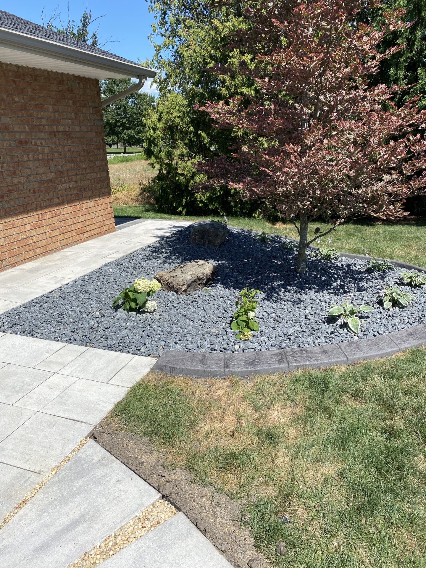 The image shows a landscaped garden with a red brick house, a stone walkway, a mulched area with young plants, a red leafy tree, and blue-gray stones.