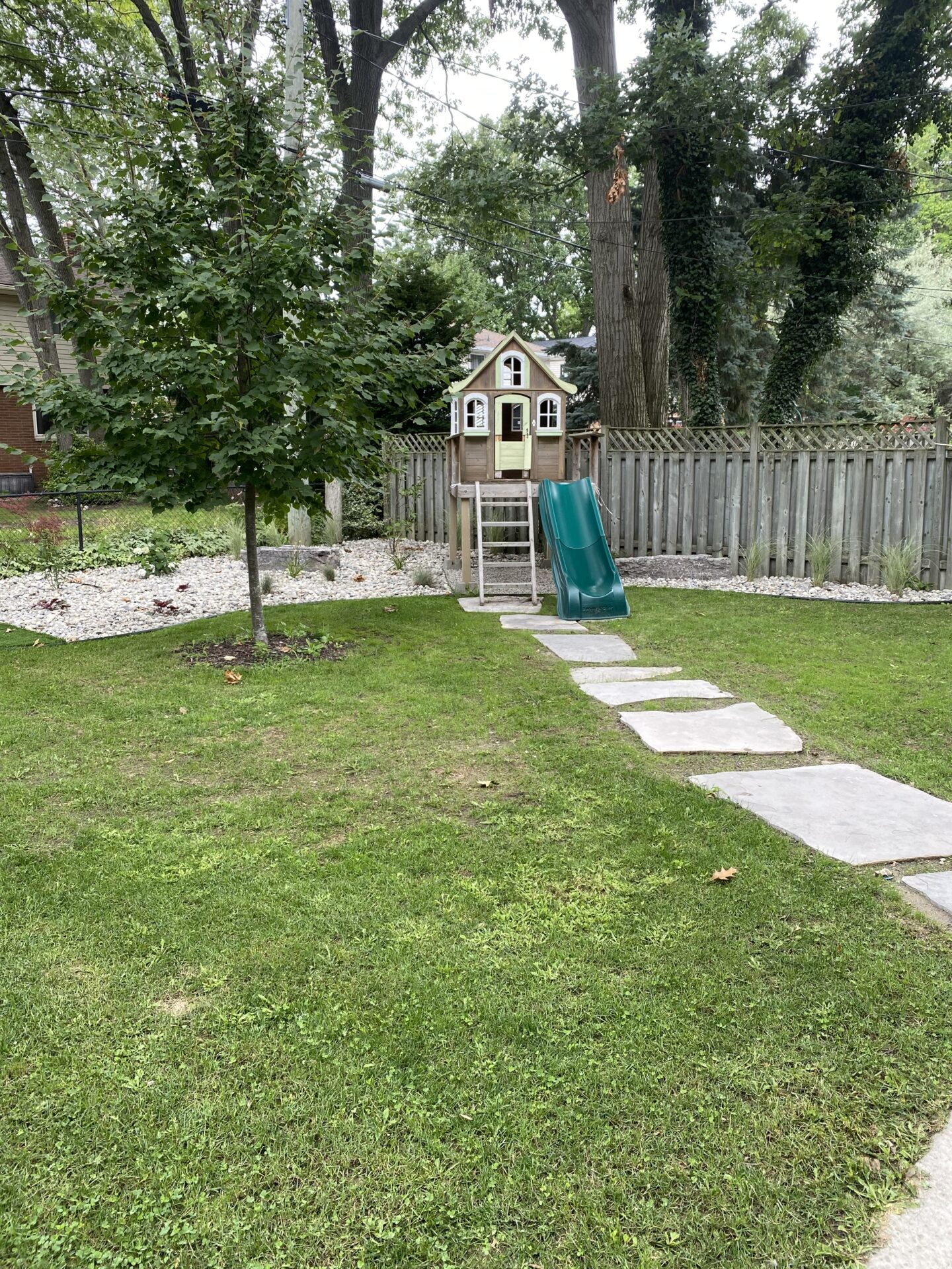 A backyard with a miniature playhouse on stilts, featuring a green slide and a stone path leading through a well-kept lawn amidst trees.
