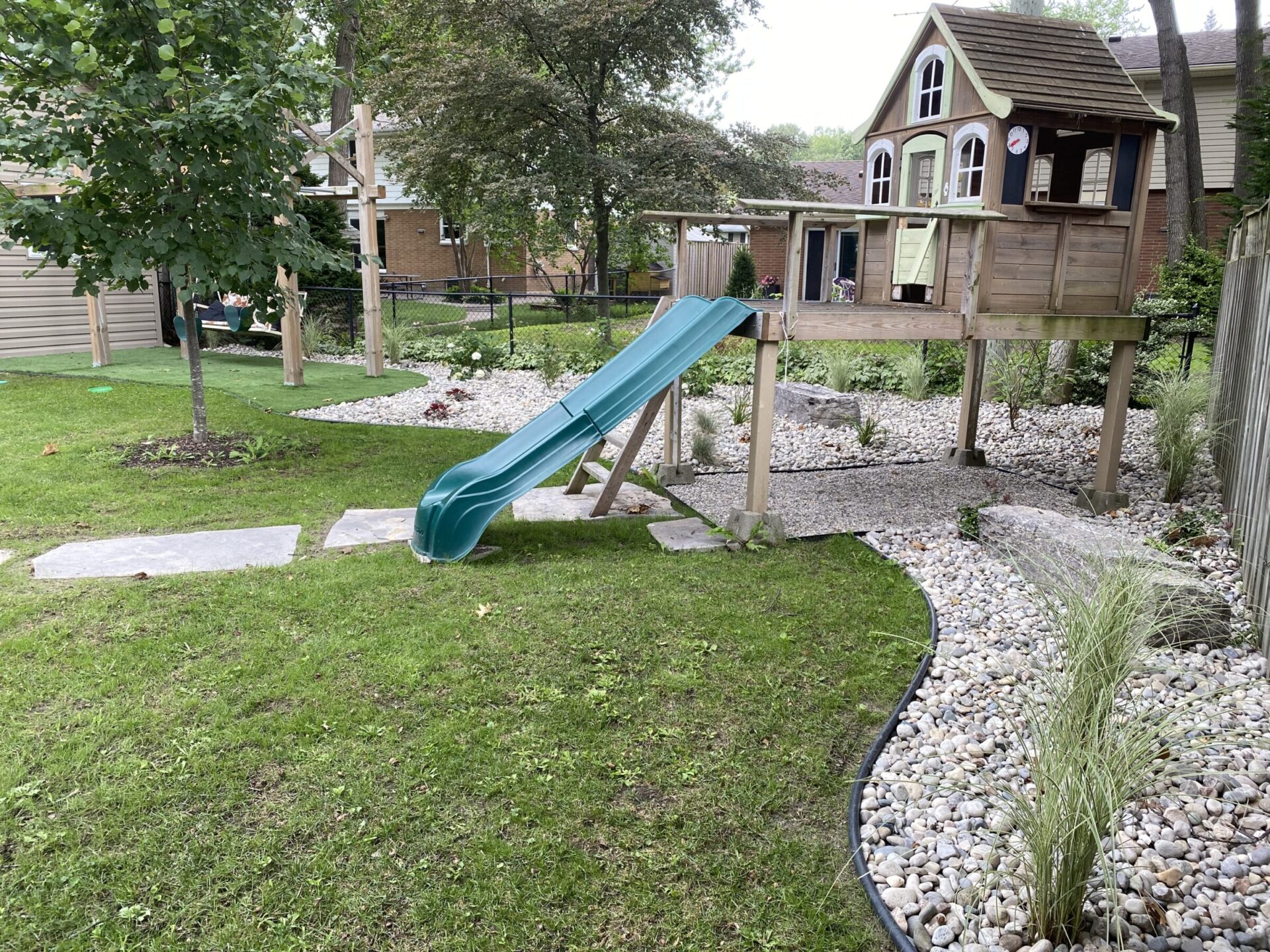A backyard playhouse on stilts with a green slide, surrounded by a landscaped yard with stepping stones, green grass, and decorative gravel.