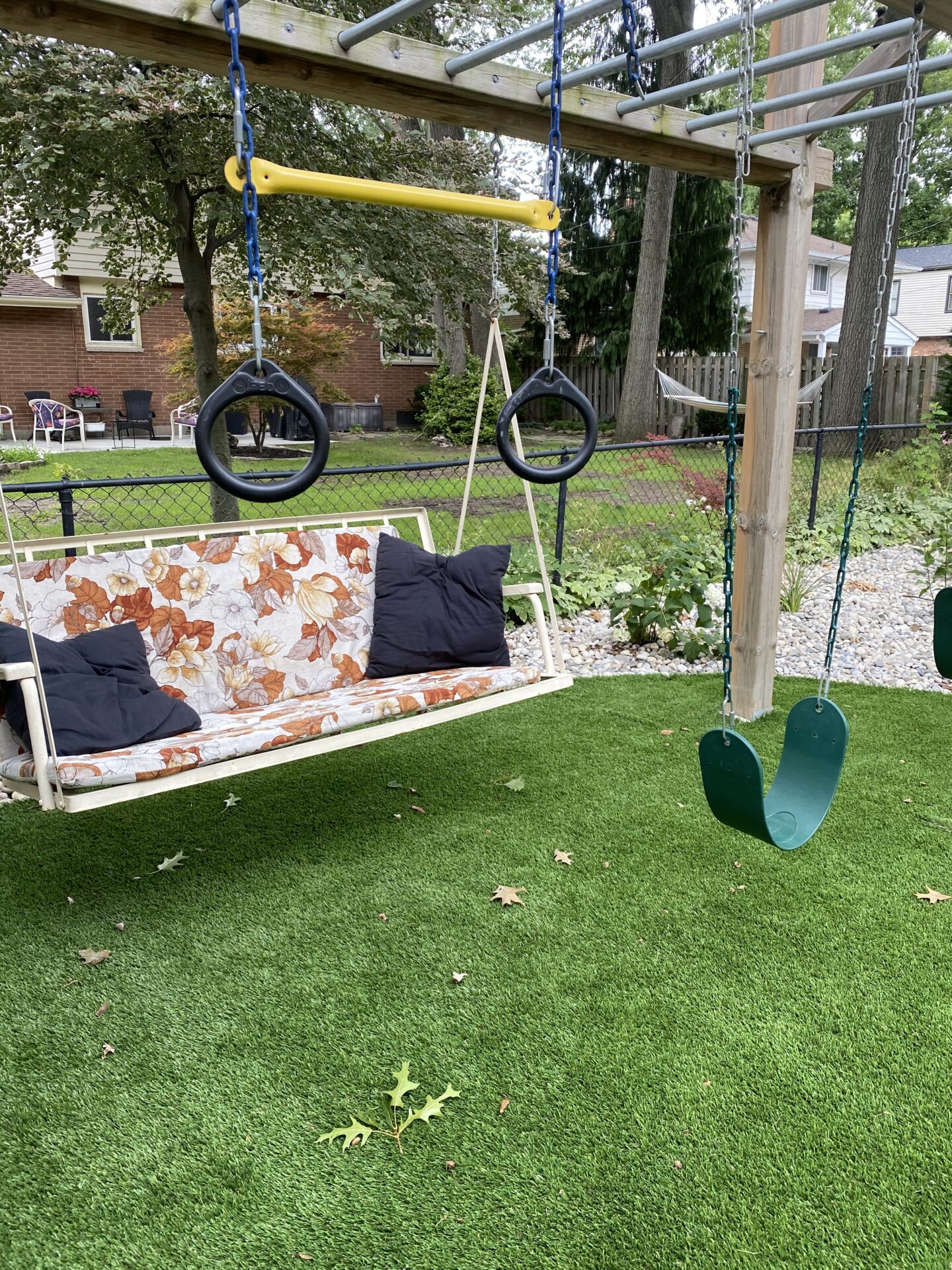 A backyard playset with rings, a swing, and a bench swing over artificial grass, scattered with leaves, adjacent to a pebble path and lawn.