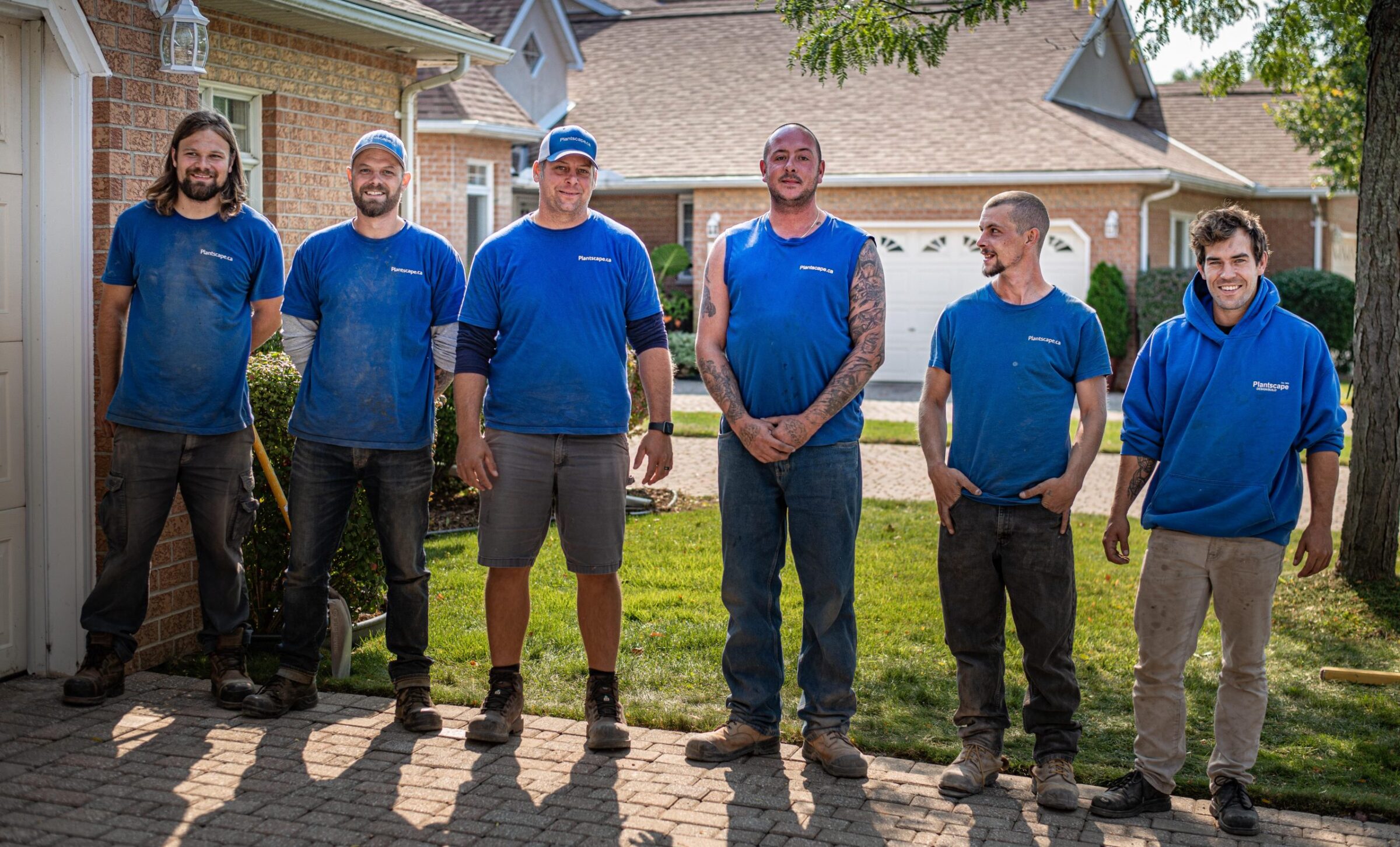 Six people in blue work shirts and jeans stand smiling in front of a residential house, with greenery and sunlight casting shadows on a driveway.