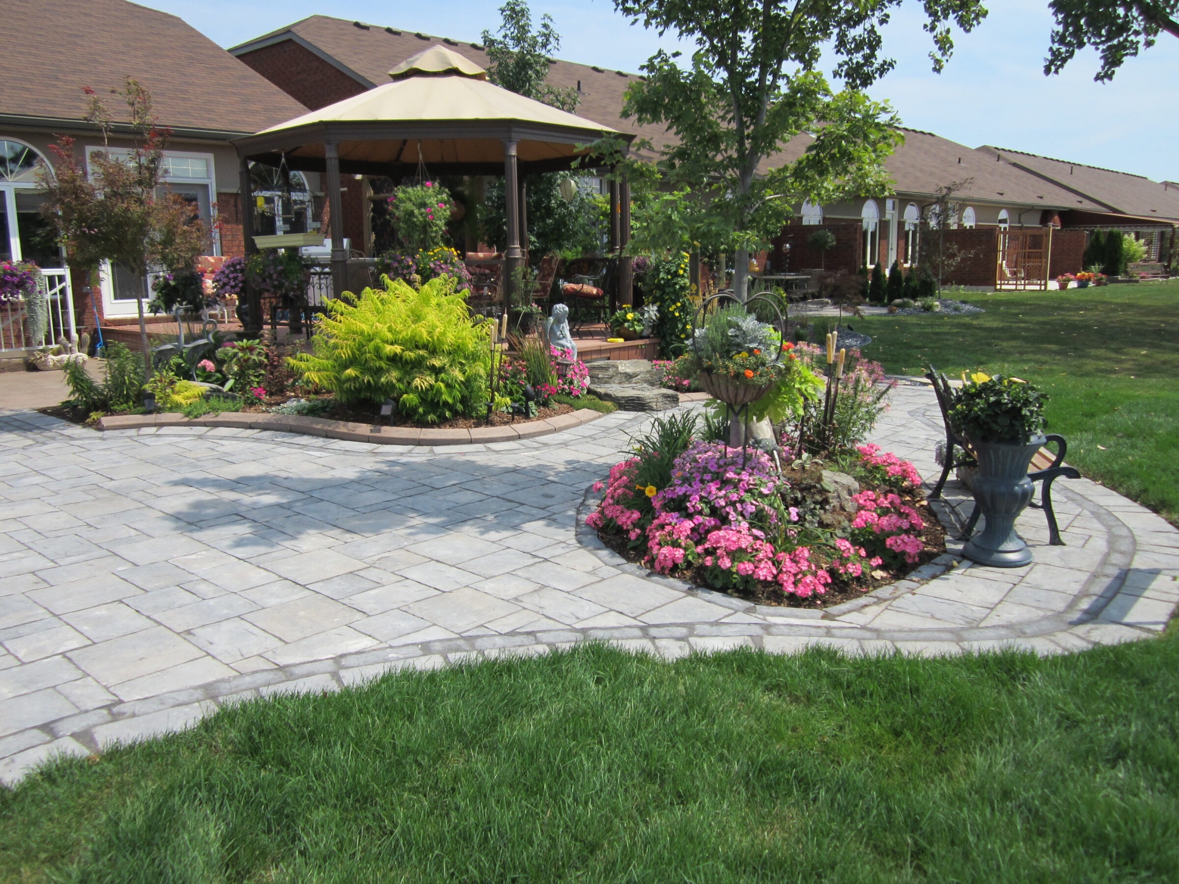This image shows a well-maintained garden with colorful flowers, a stone pathway, a patio area with a sunshade, green lawn, and a suburban house.