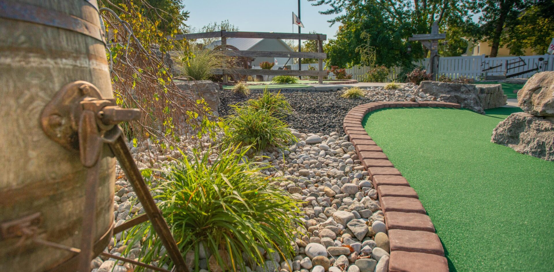 This image shows an outdoor mini-golf course with green artificial turf, edged by brick trim, surrounded by pebbles, rocks, and plants on a sunny day.