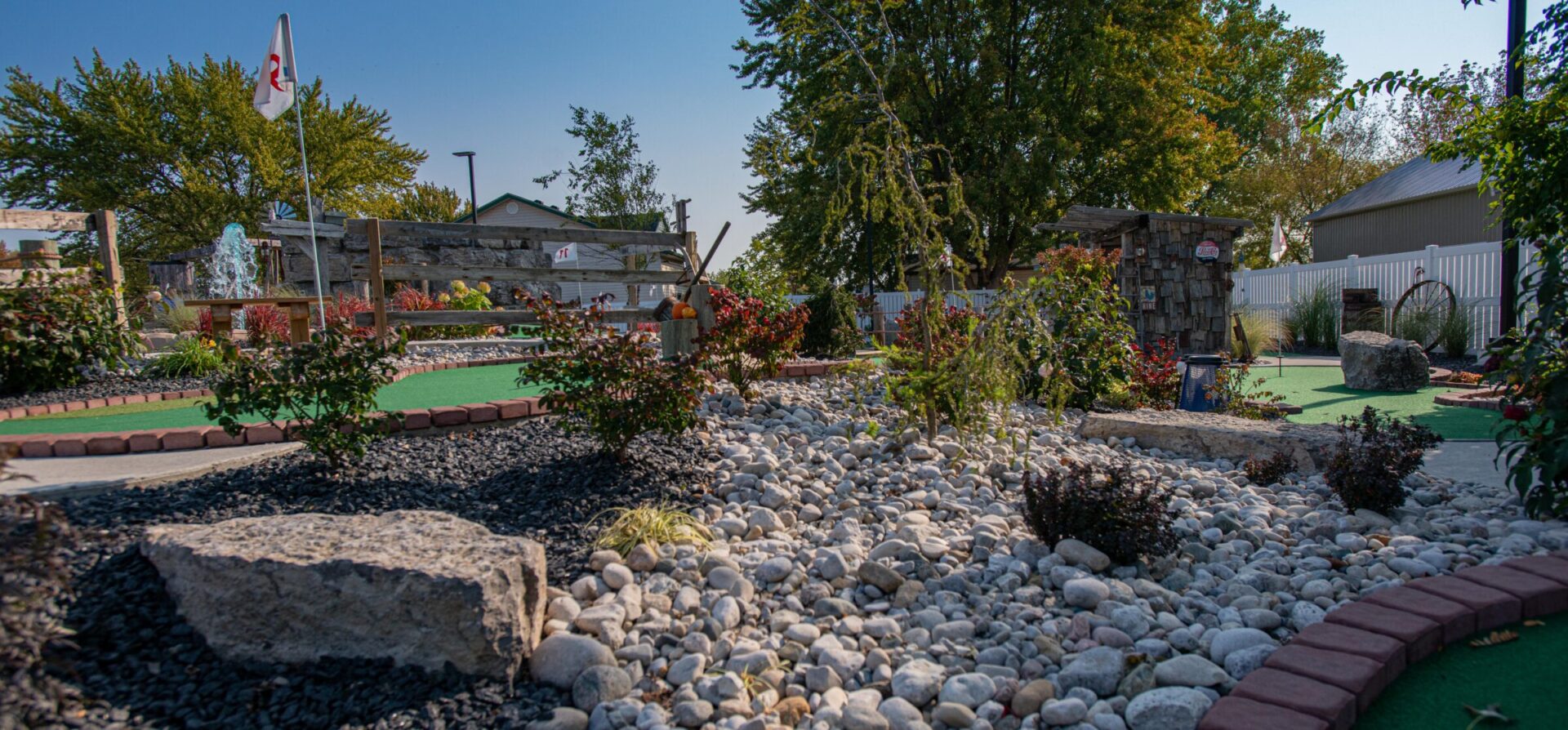 An outdoor miniature golf course featuring green turf, decorative rocks, plants, a small fountain, and a clear blue sky above.