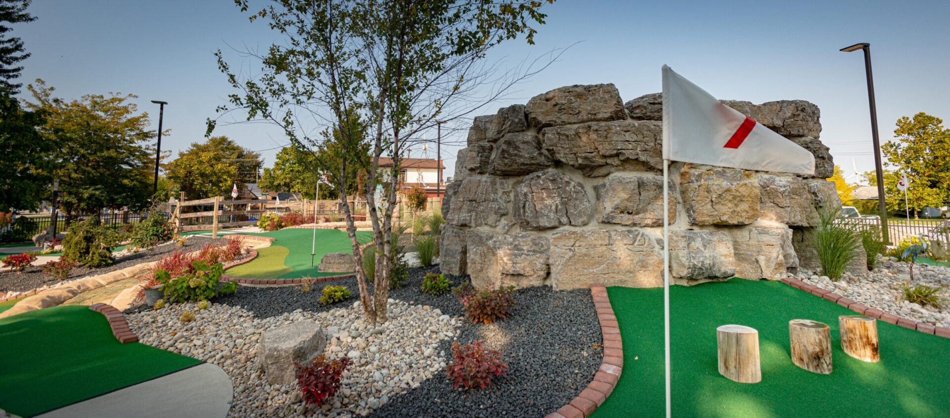 A mini-golf course with green artificial turf, a flag on a hole, decorative rocks, and landscaping under a clear blue sky.
