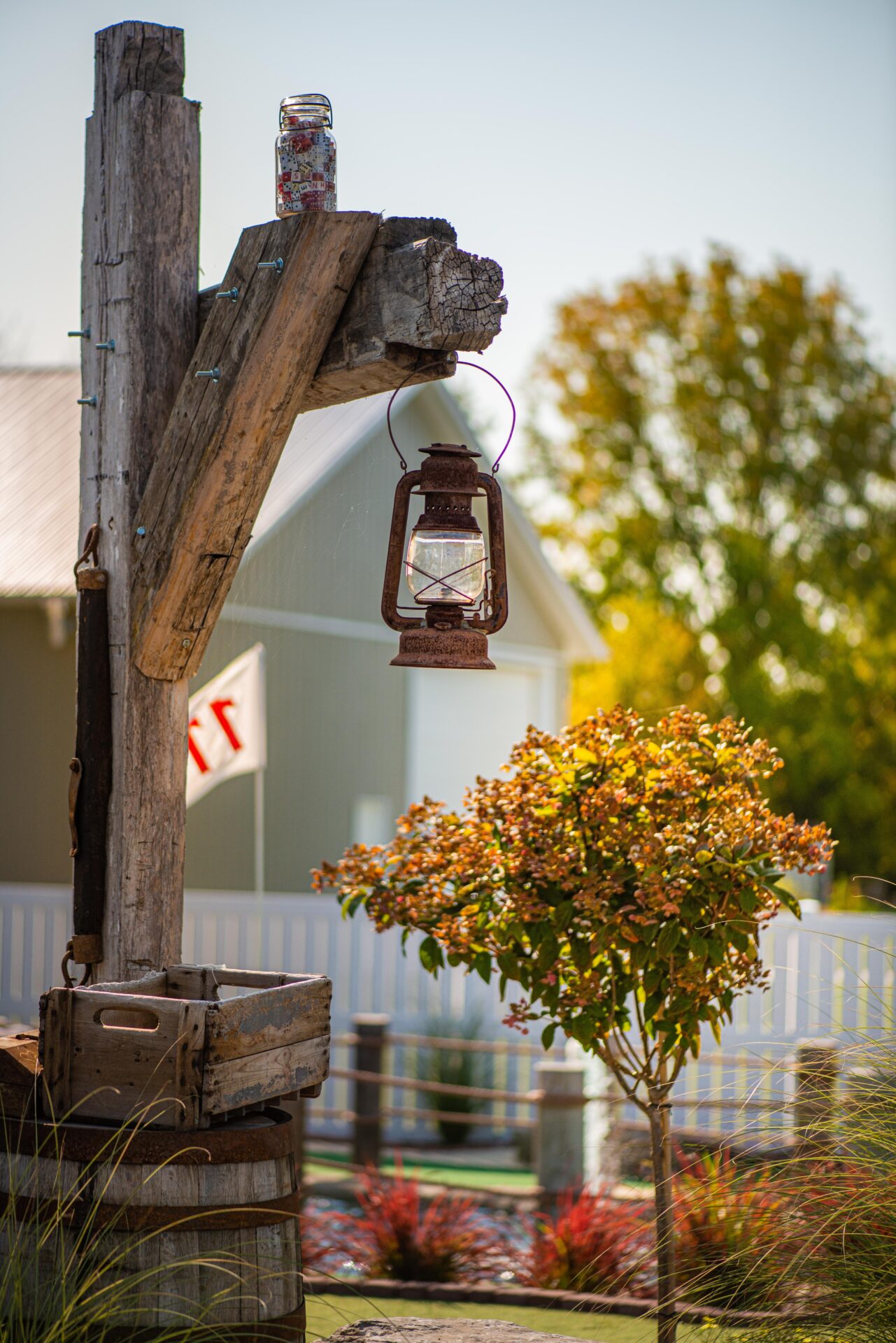 An old rustic lantern hangs from a weathered wooden post with jars on top, beside a flowering bush, with a white picket fence background.
