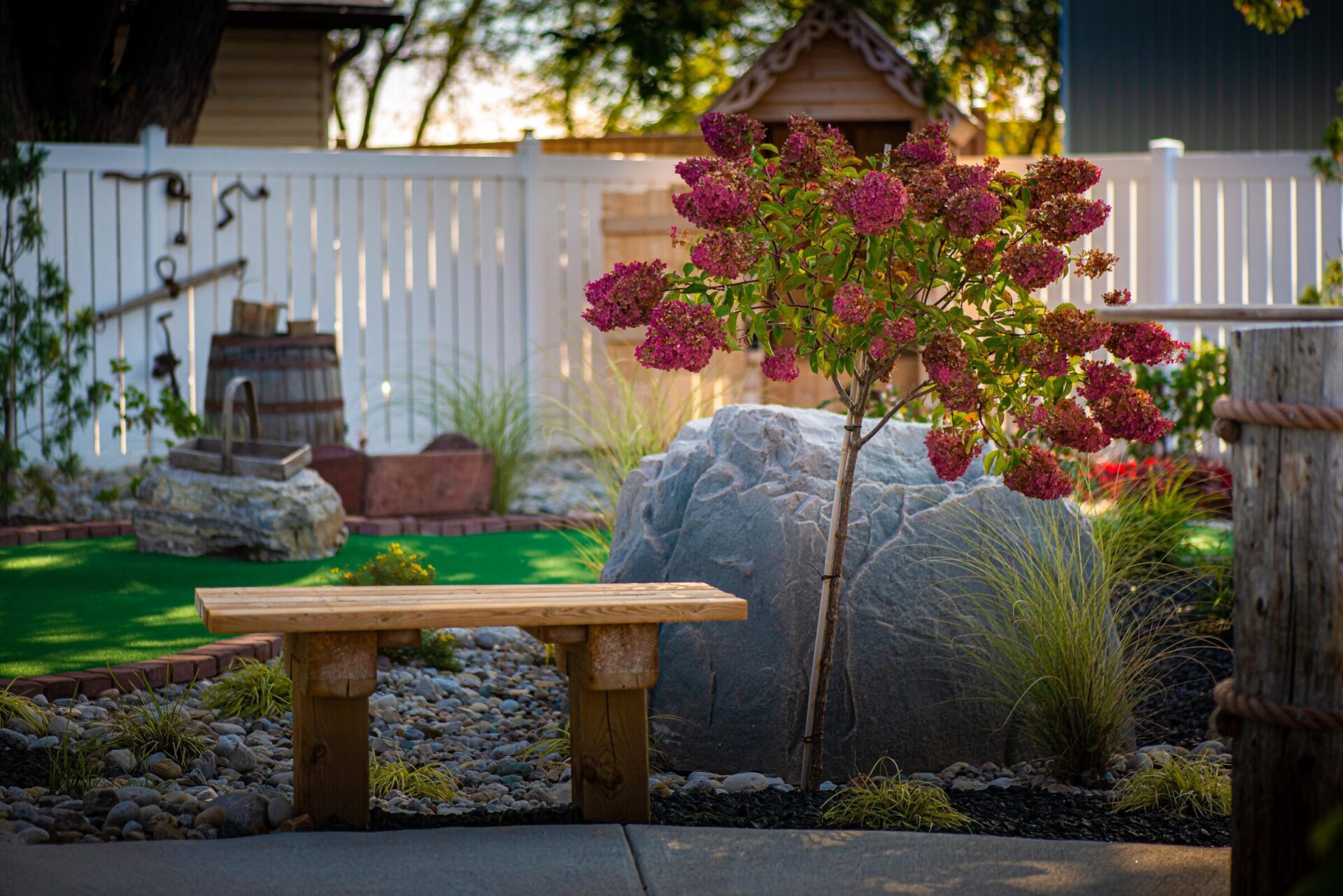 The image features a serene garden with a flowering shrub, a wooden bench, rocks, grasses, a miniature windmill, and a white picket fence.