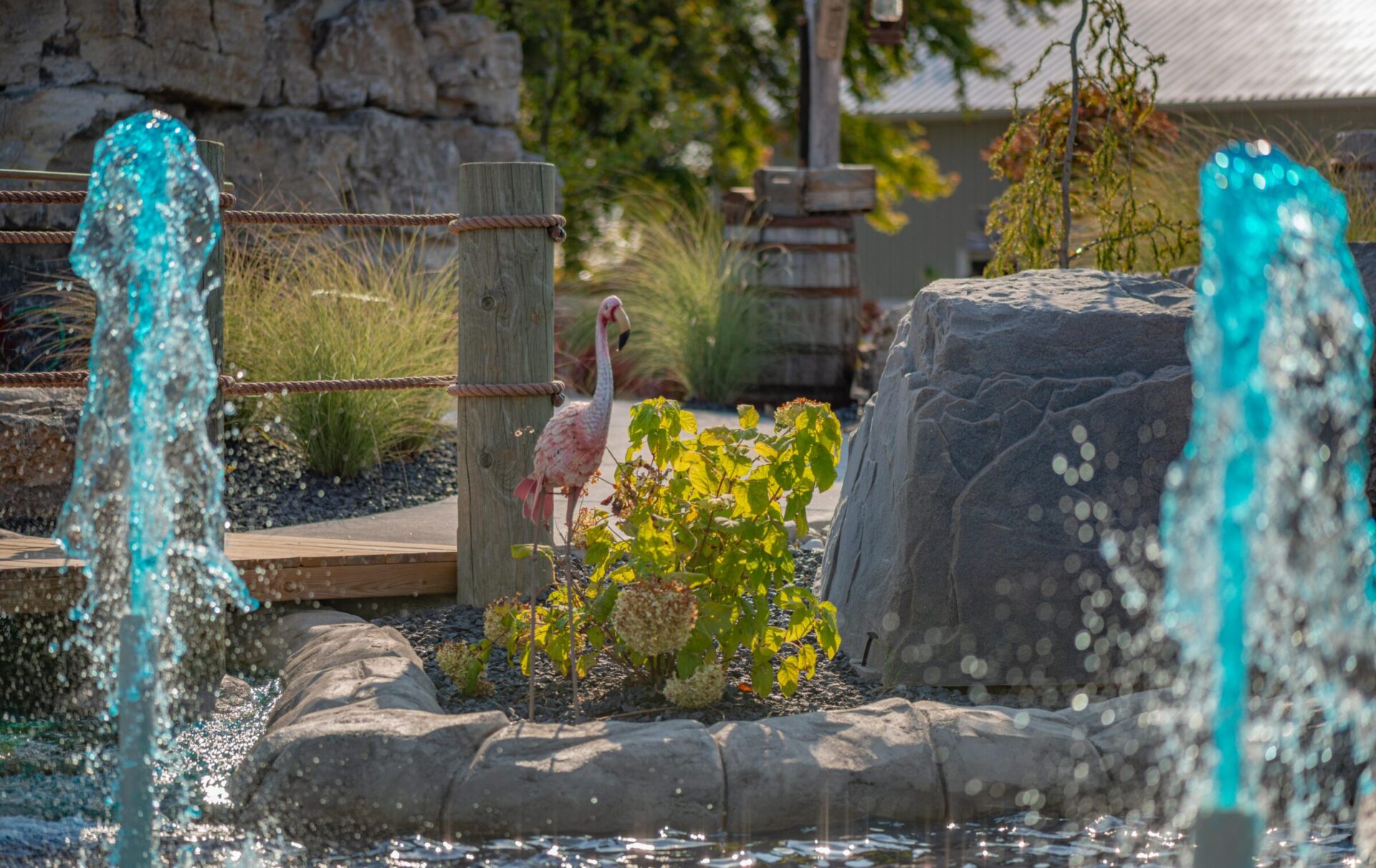 The image shows a vibrant garden setting with water jets, rocks, and a flamingo sculpture amidst green foliage, under a clear, sunny sky.