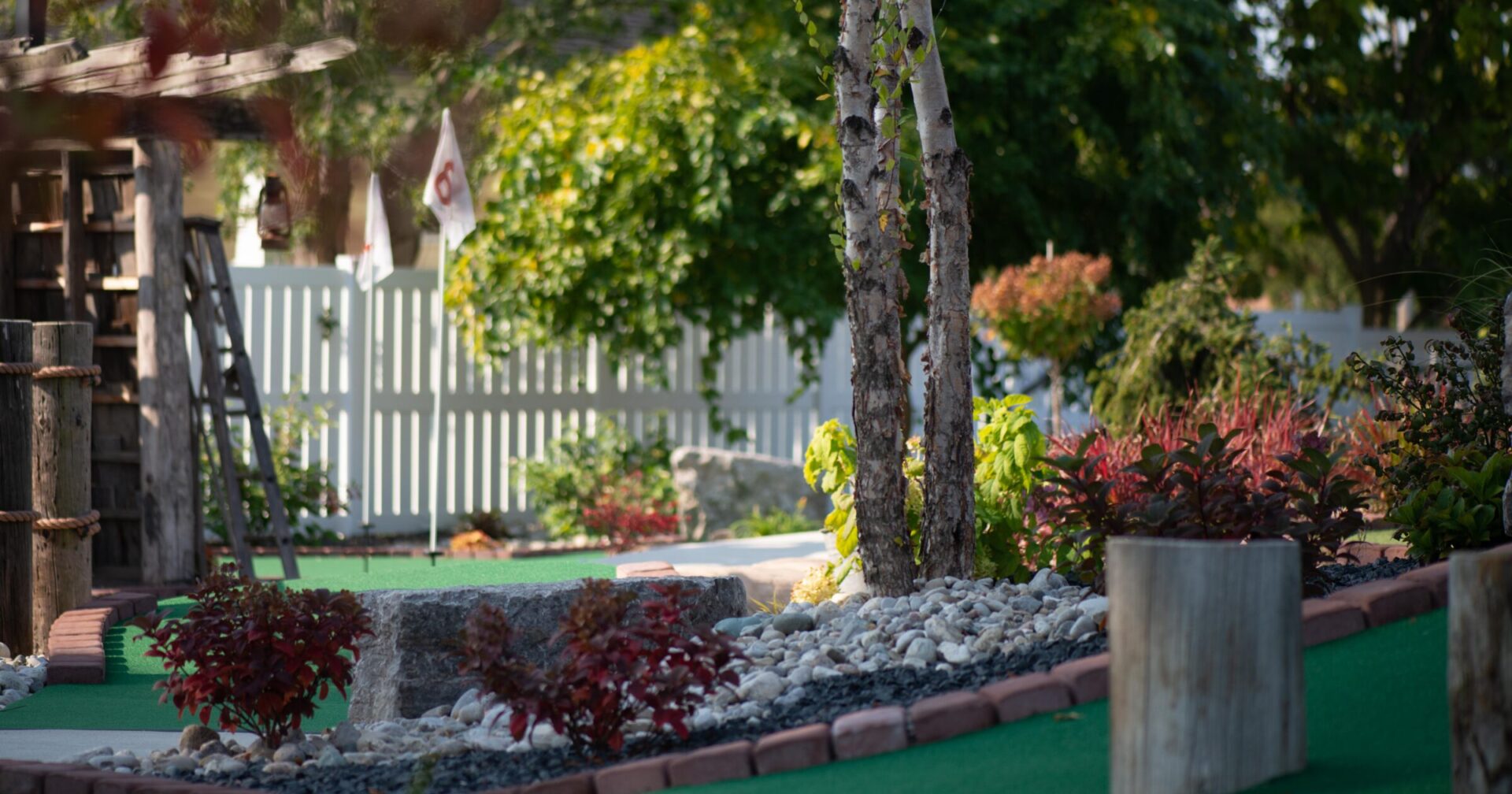 A tranquil mini-golf course with green artificial turf, surrounded by decorative shrubs, birch trees, white fencing, and a rustic wooden structure.