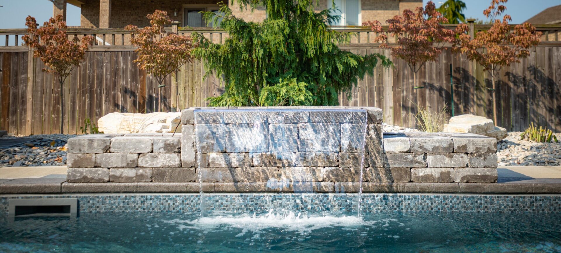 This image features an outdoor swimming pool with a stone waterfall feature, surrounded by a wooden fence and landscaping under a clear sky.