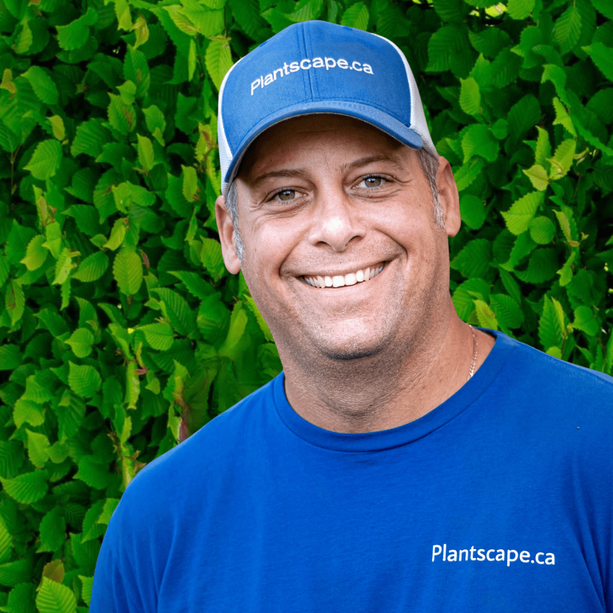 A person with a friendly smile, wearing a blue cap and t-shirt branded 'Plantscape.ca,' stands in front of a lush green leafy background.