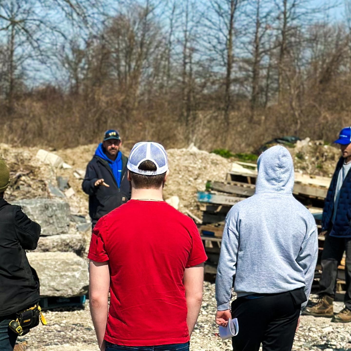 A group of people, with one gesturing and explaining, are standing outdoors in a construction area with concrete debris and bare trees in the background.