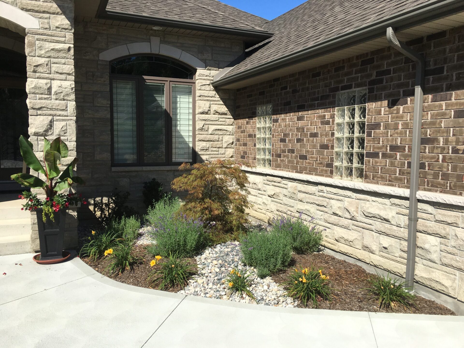 This image shows a well-maintained front yard with a stone exterior house, landscaped with shrubs, flowering plants, and a decorative potted plant.