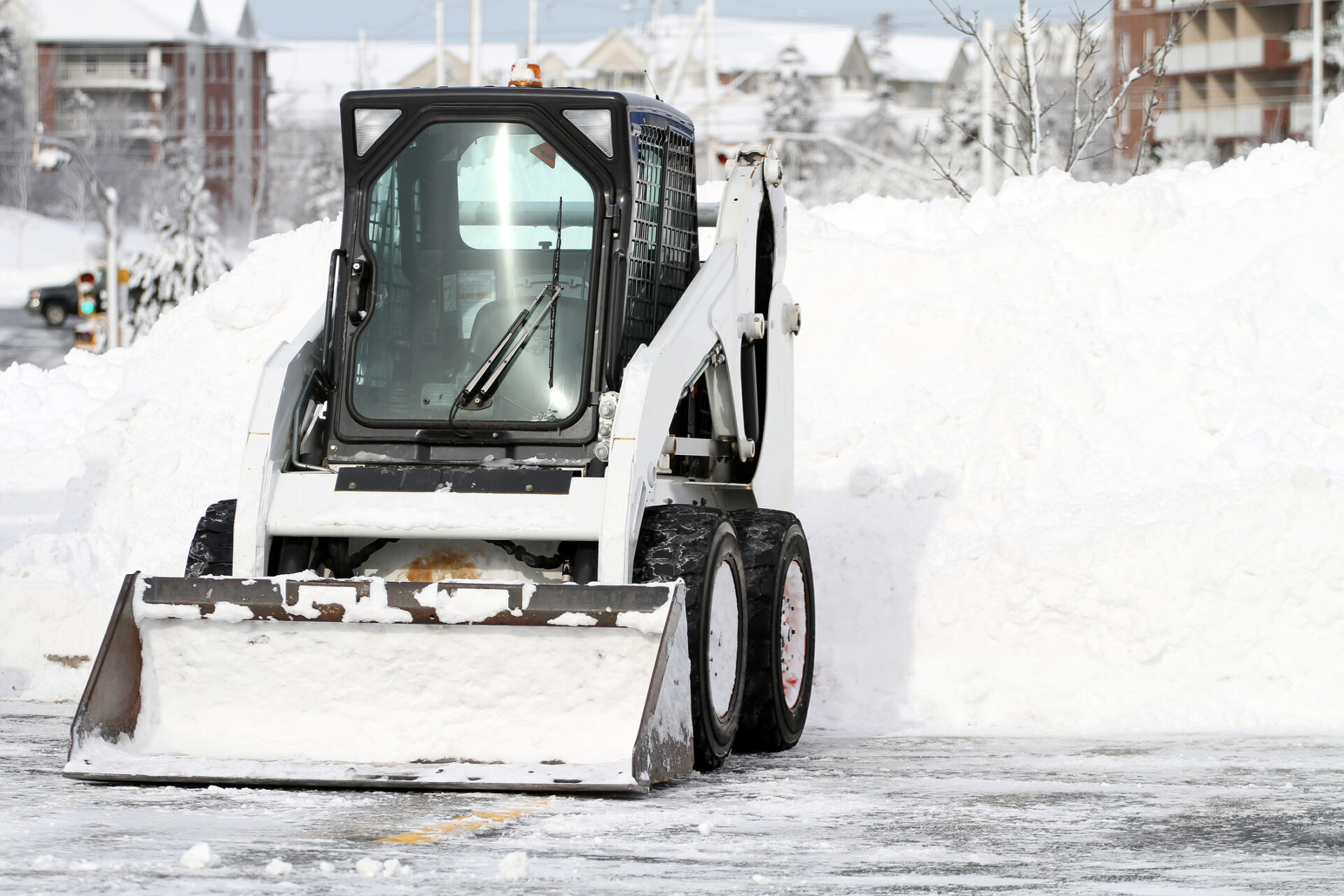 A skid-steer loader is clearing snow in a parking lot with mounds of plowed snow visible and winter buildings in the background under a bright sky.