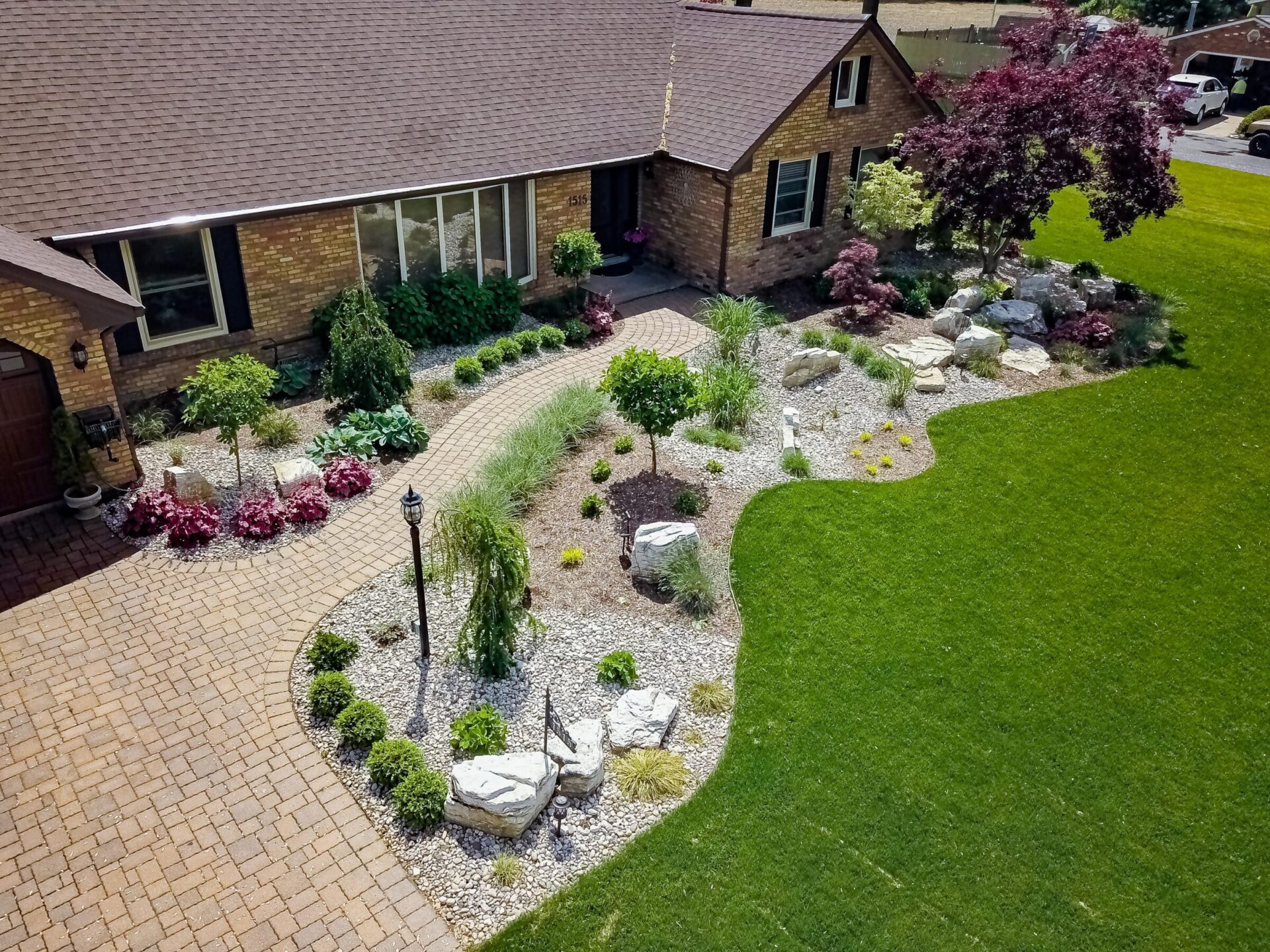 Aerial view of a landscaped front yard with a brick house, curved pathways, lush lawn, assorted plants, decorative rocks, and a lamppost.