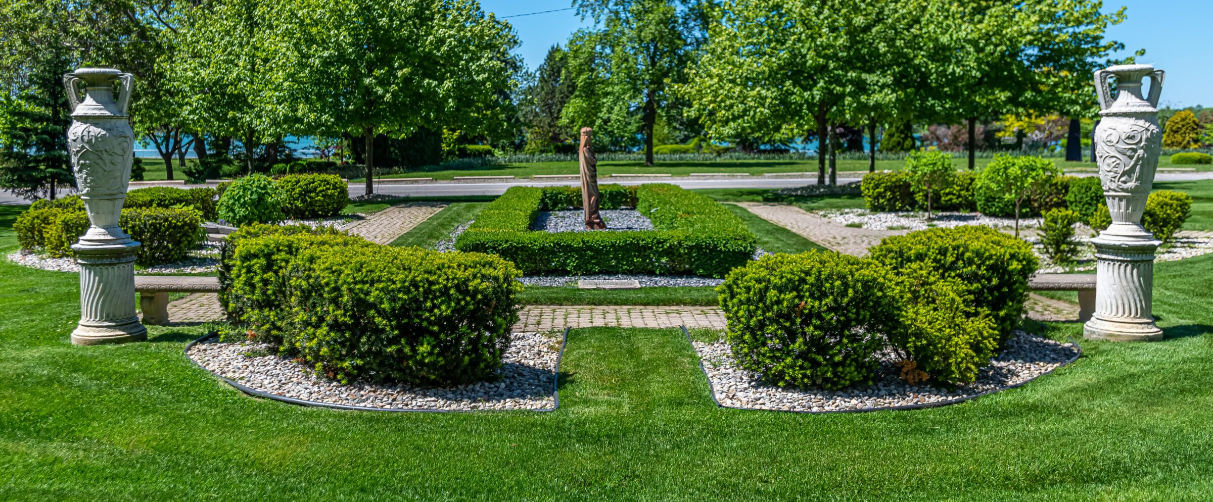 This image shows a symmetrical formal garden with sculpted shrubs, ornate urns, brick pathways, a central statue, under a clear blue sky.