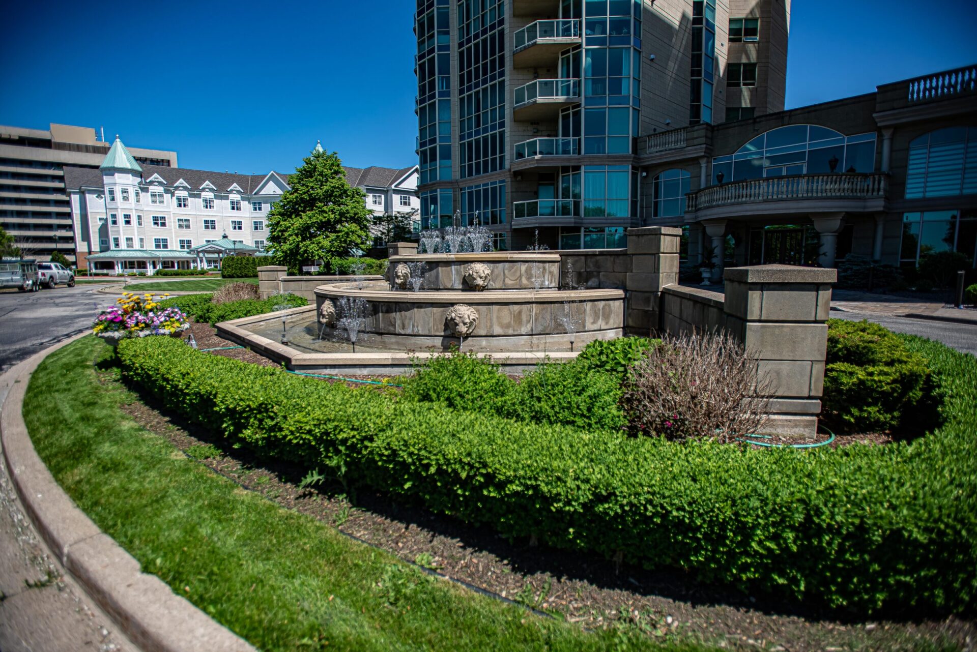 The image shows a vibrant outdoor setting with a fountain, manicured hedges, and a variety of buildings under a clear blue sky.