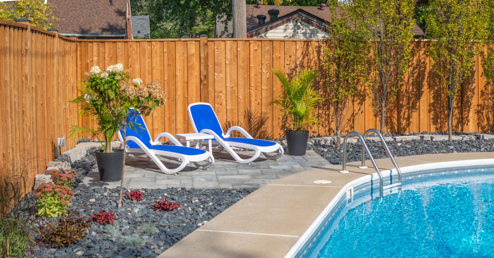 This image shows a serene backyard setting with a swimming pool, blue lounge chairs, potted plants, a stone path, and a wooden fence.