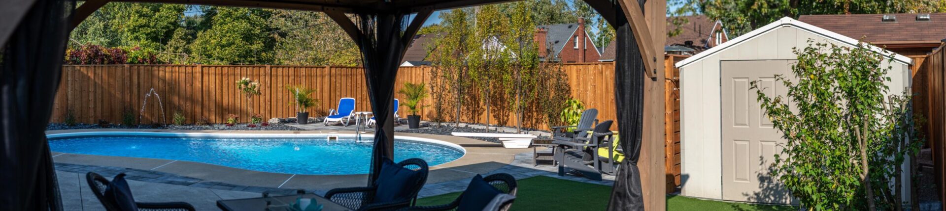 Panoramic view from a shaded patio of a sunlit backyard with an inviting swimming pool, lounge chairs, a neat fence, and a storage shed.