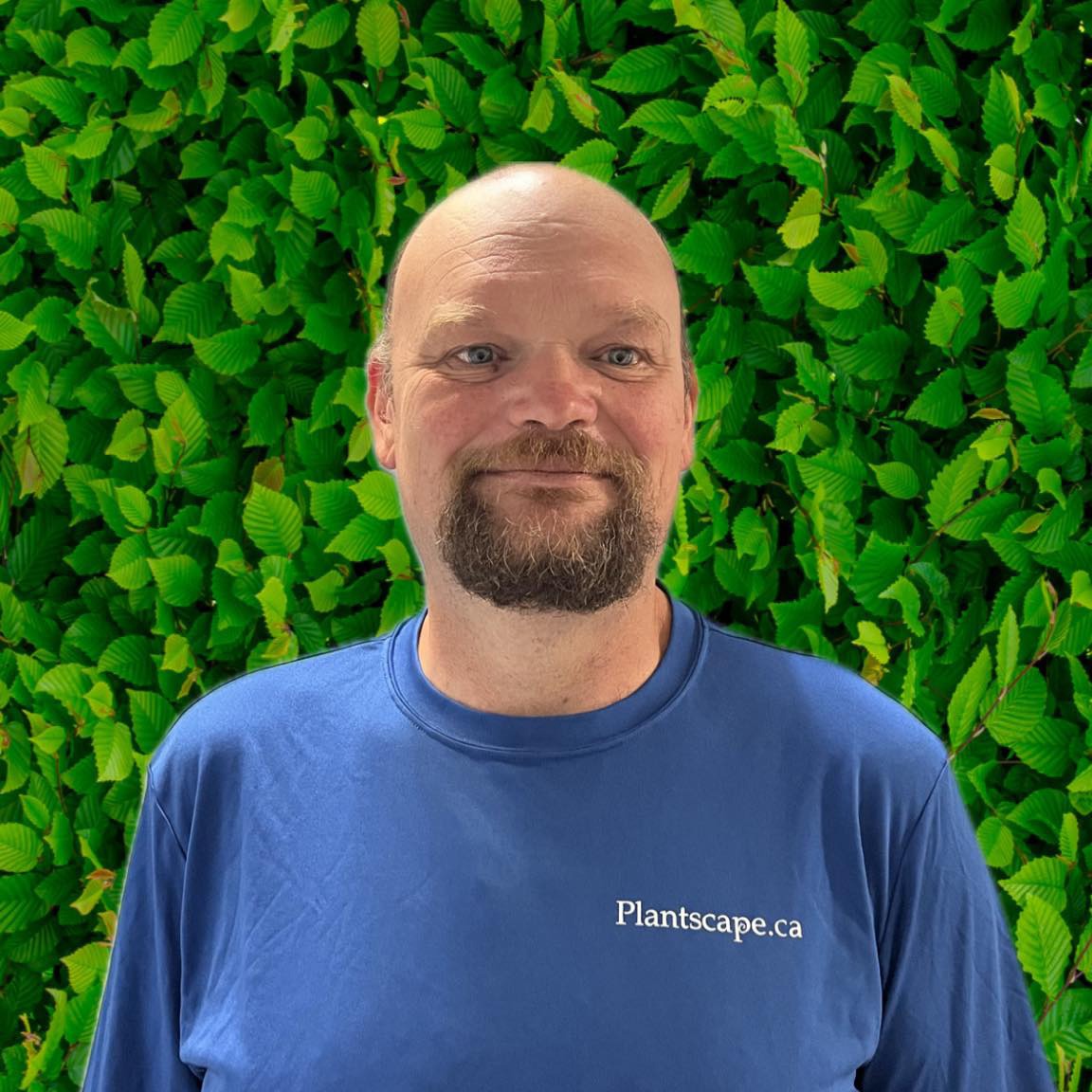 A person with a beard is smiling in front of a lush green leafy background, wearing a blue shirt with the text "Plantscape.ca" on it.